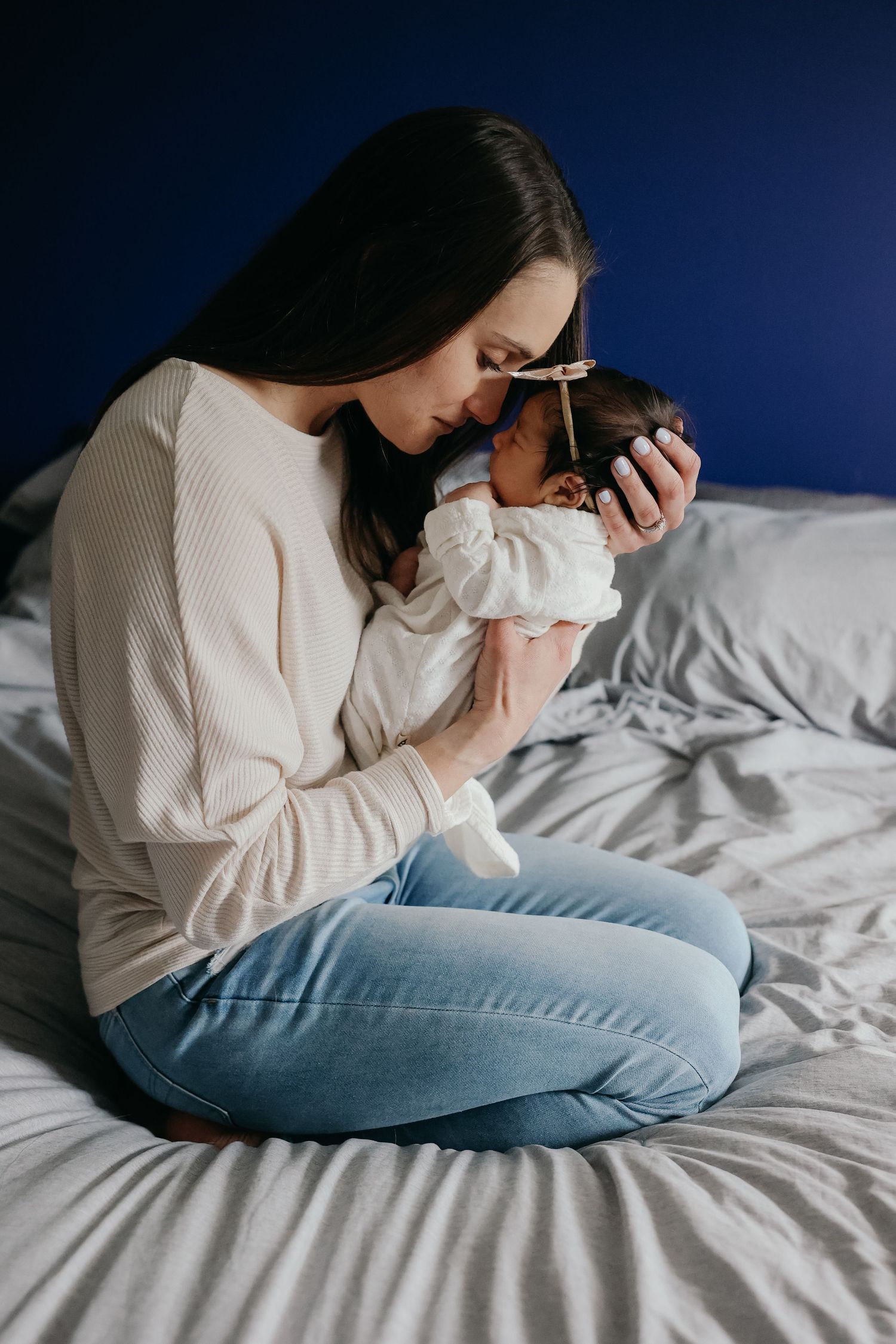 Person in cream sweater sitting on bed holding and nuzzling a newborn baby dressed in white.