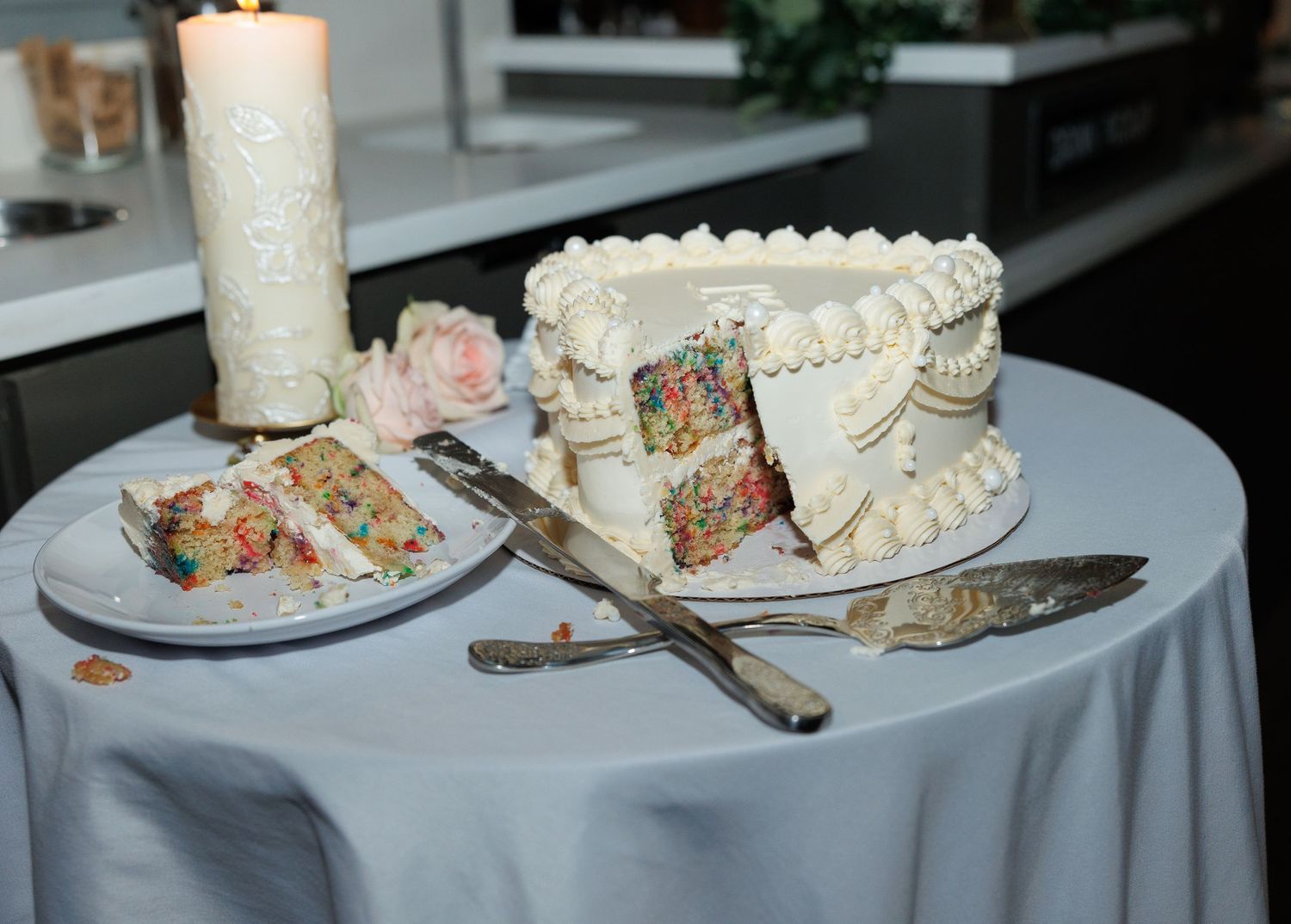 White frosted funfetti wedding cake with sliced piece on plate, cake knife, and candle on gray tablecloth.