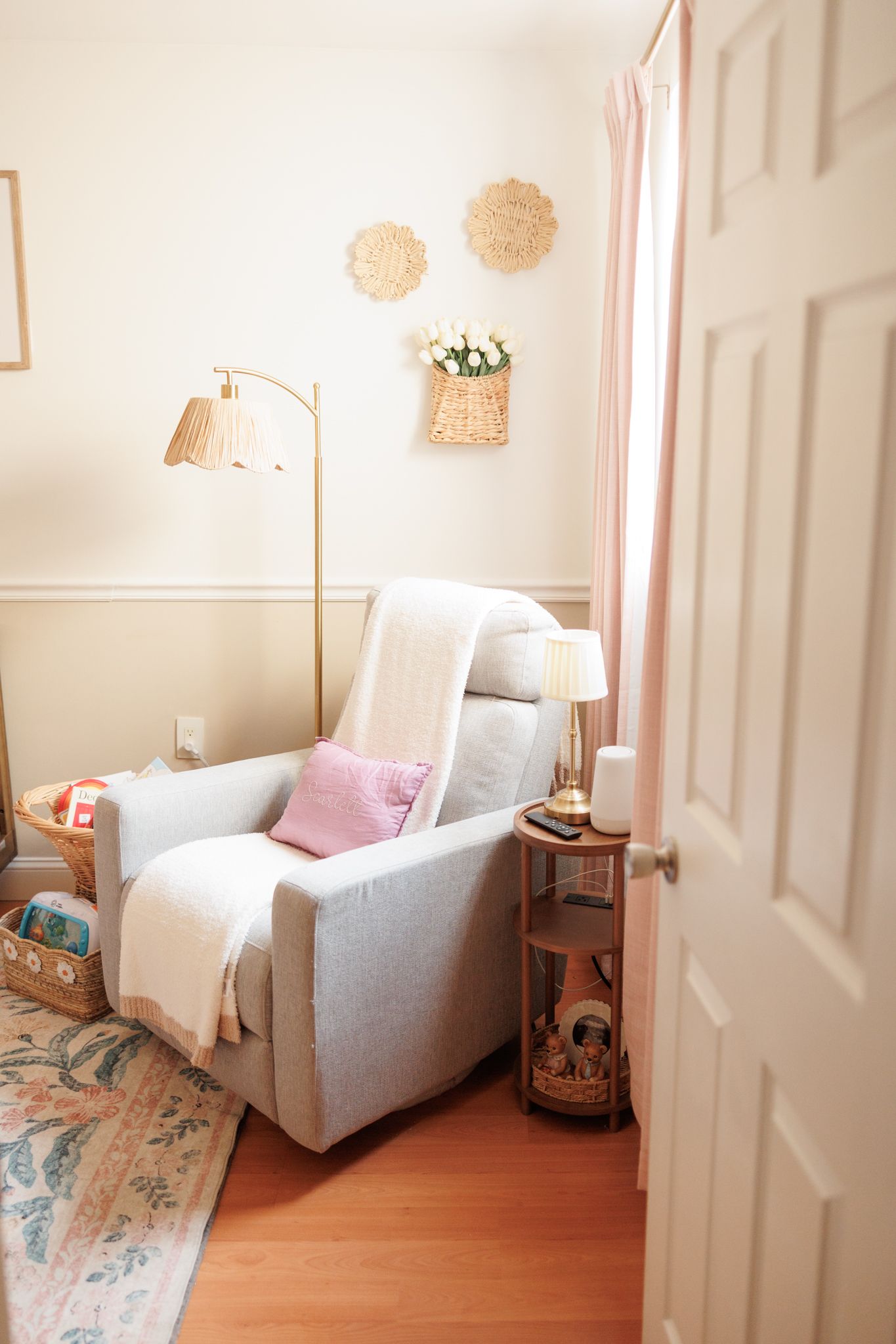 Cozy nursery reading nook with gray glider chair, pink cushion, white throw, floor lamp, and wicker wall decor.