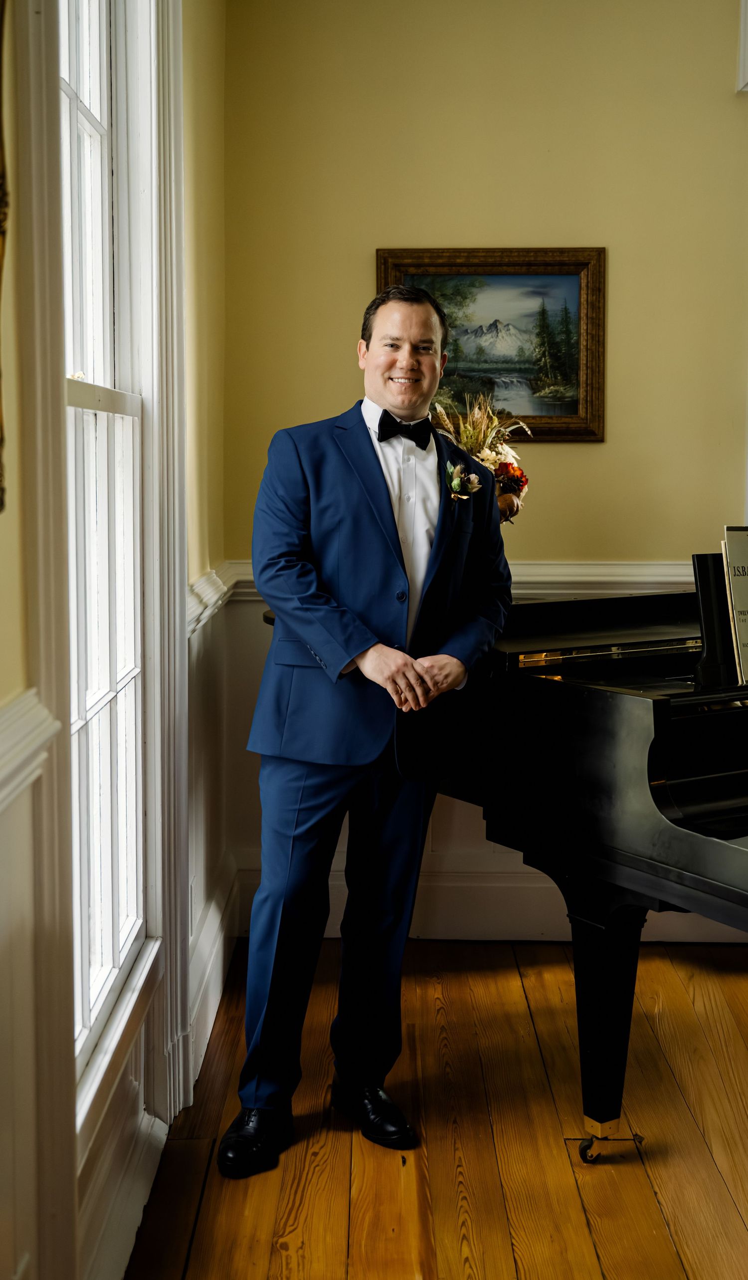 Groom in navy tuxedo with floral boutonniere poses beside a grand piano near a window in farm house at Guildford Farm in Ruckersville, VA.