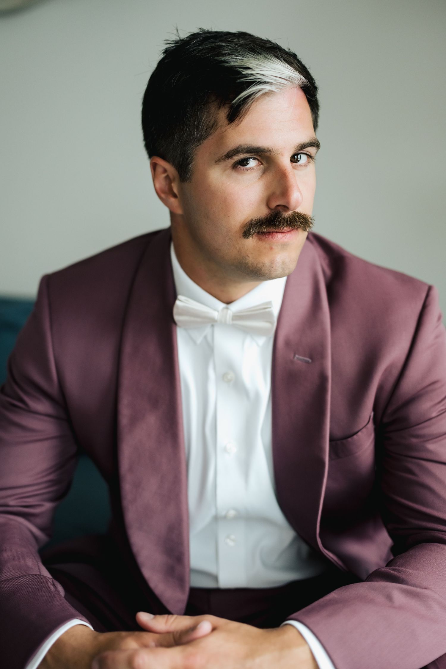 Dapper groom in burgundy tuxedo with white bow tie poses for portrait.