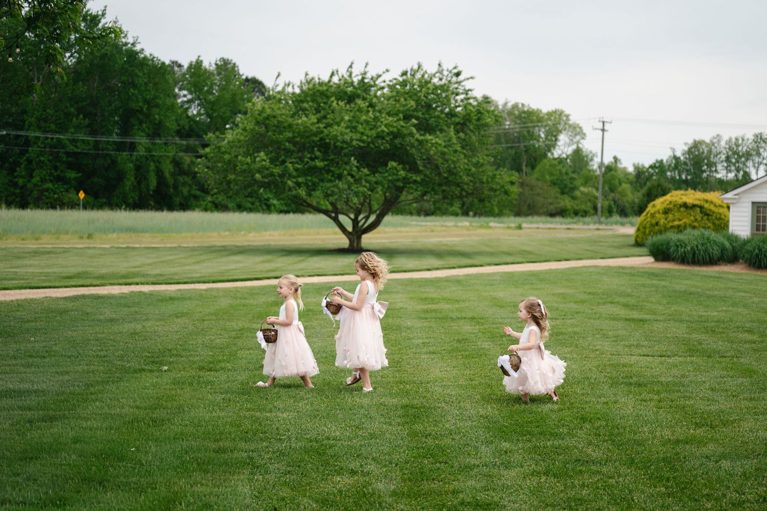 Three flower girls in white dresses playfully spinning on a lush green lawn at a wedding at The Barns at Timberneck in Smithfield VA.