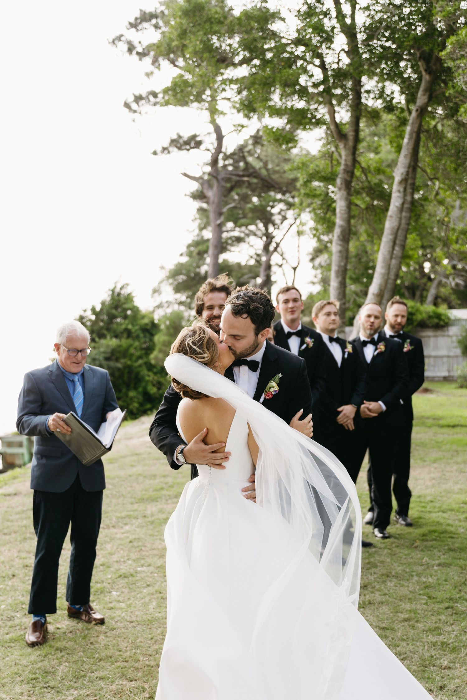 Groom dips bride in white gown for a kiss during outdoor wedding ceremony with groomsmen behind at The Westside Inn in Manteo, NC.