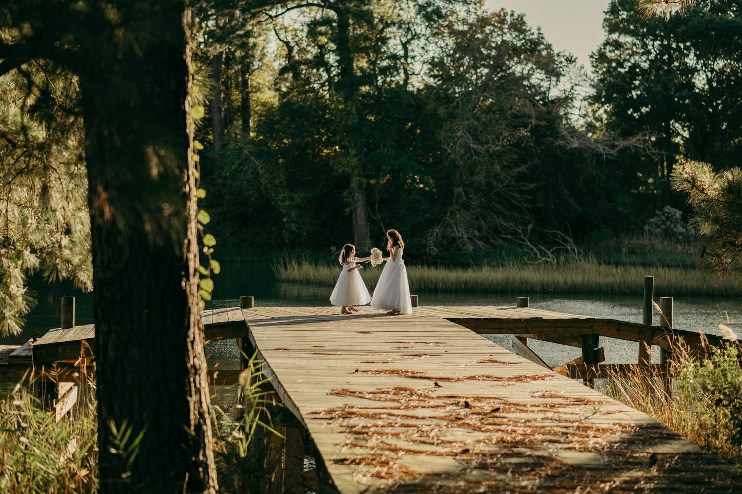 Two flower girls in white dresses hold hands on a wooden dock surrounded by a peaceful lake and forest landscape during a family estate wedd