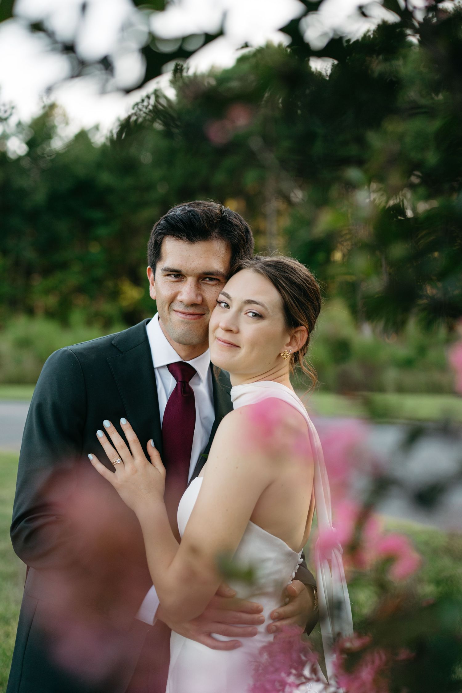 Bride in white halter gown and groom in dark suit embrace among pink blooms at the Main of Williamsburg Wedding Venue.