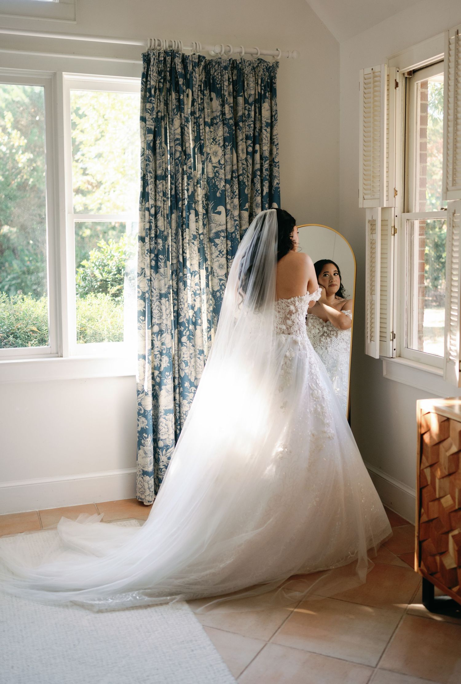 Bride in a lace gown and long veil gazes into a mirror while getting ready for her wedding at Church Point Manor in Virginia Beach.