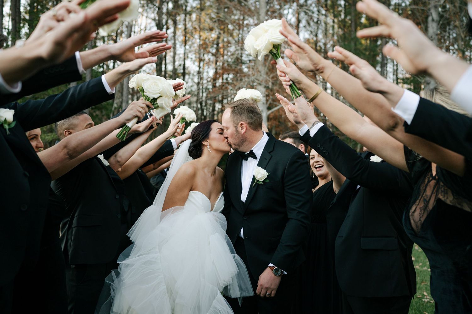 Wedding party raises bouquets to form an arch as bride and groom share a kiss during their outdoor wedding portrait at the Main at Williamsb