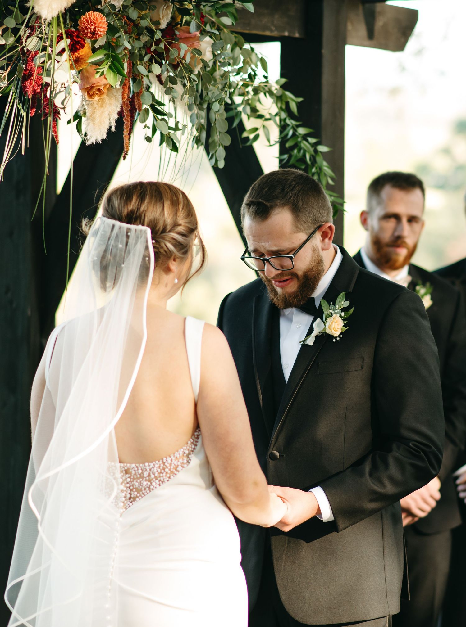 Groom in a black suit cries warmly at his bride during their outdoor wedding ceremony beneath a floral arch at Lovingston Winery in Virginia