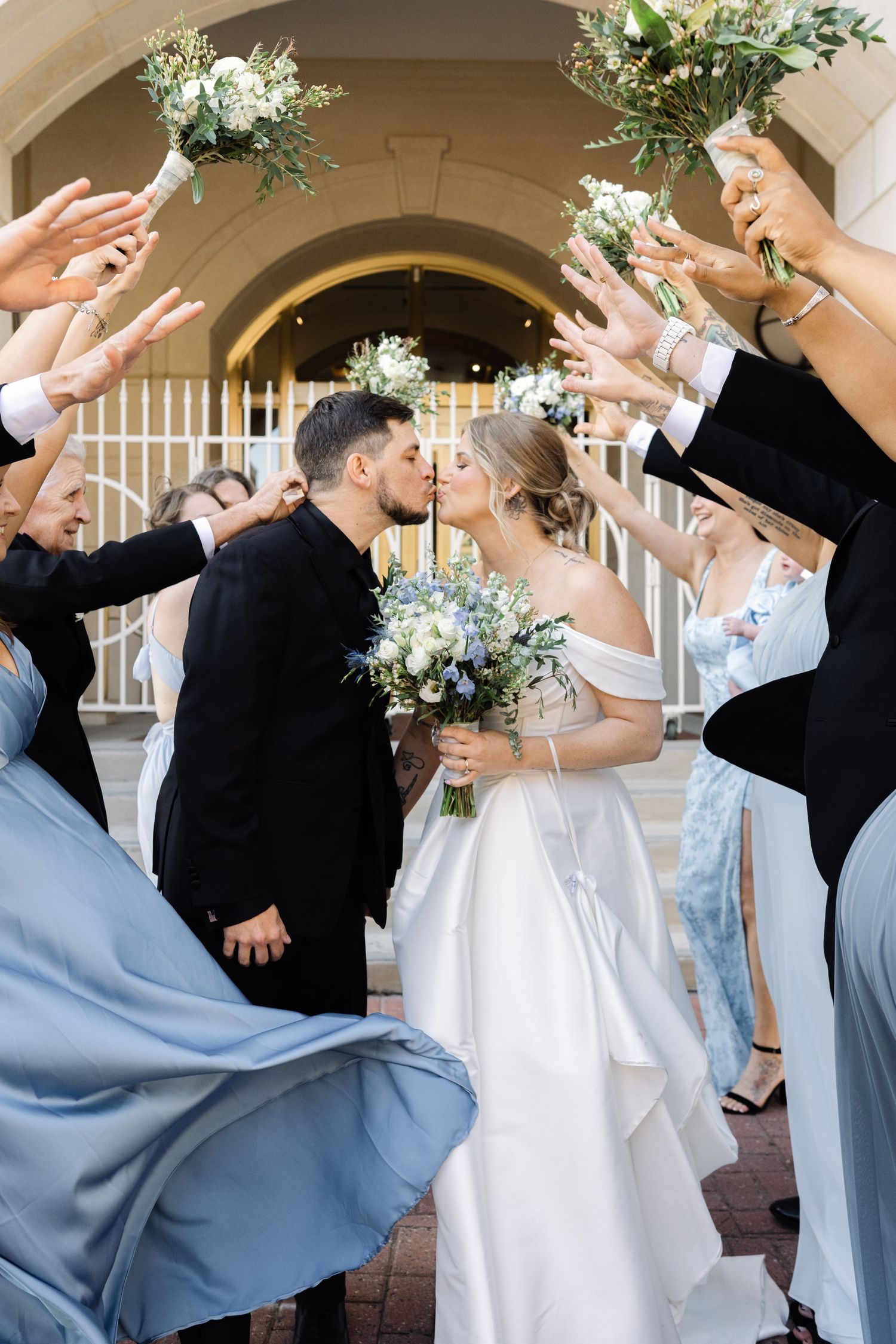 Bride and groom share kiss outside church while wedding party raises bouquets.