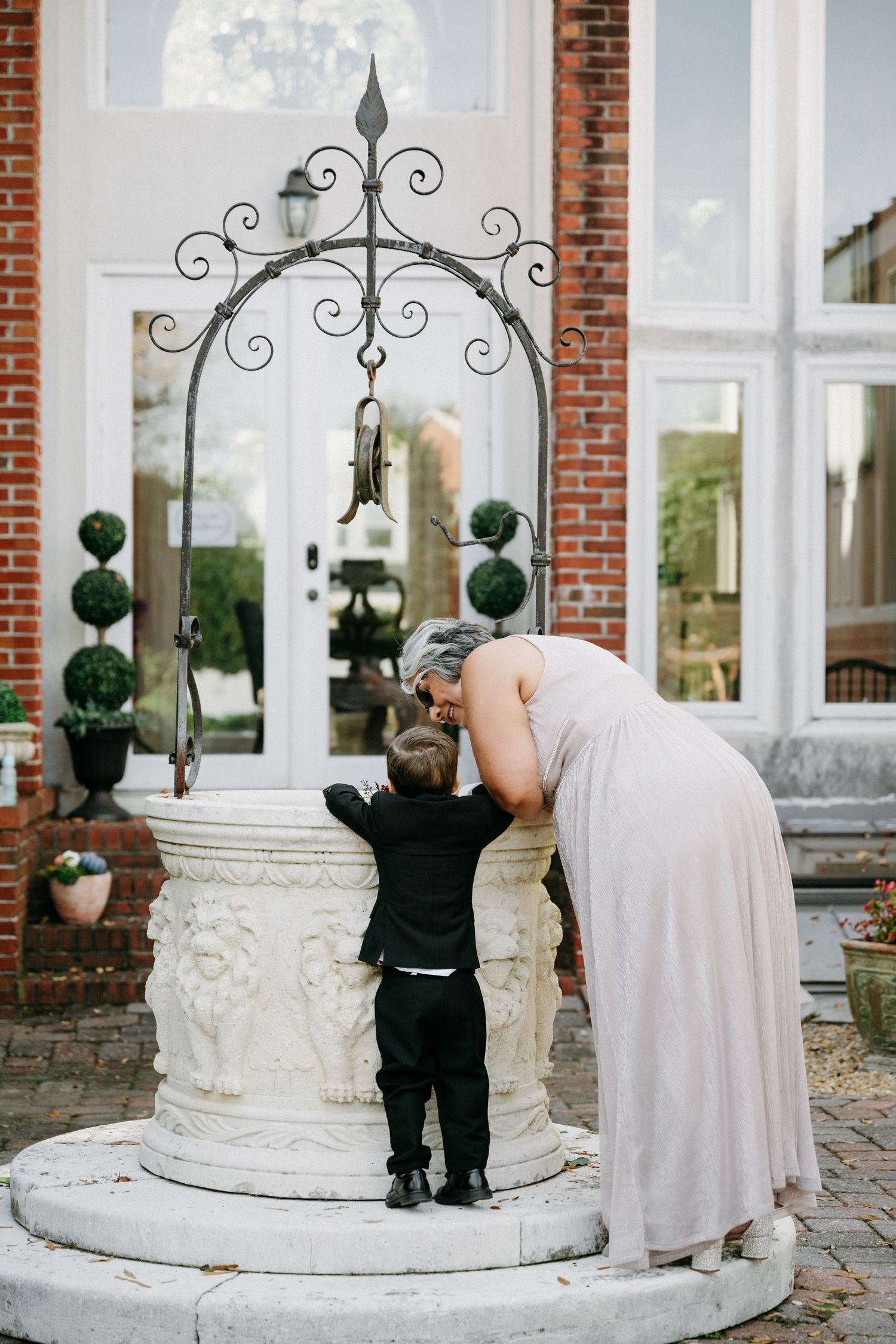 Bridesmaid leans down to a young boy in a suit at an ornate stone wishing well at Church Point Manor wedding in Virginia Beach.