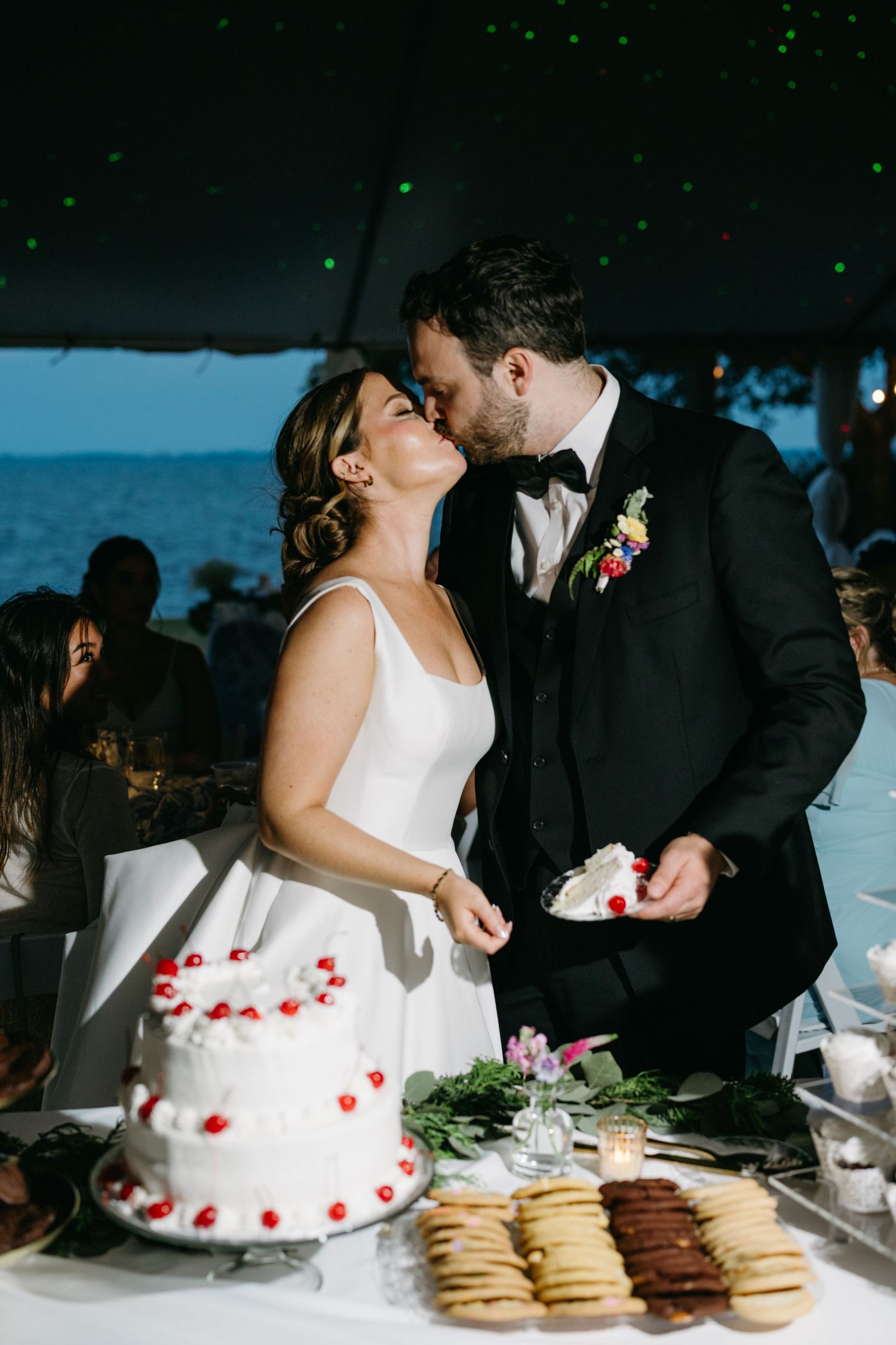 Bride and groom kiss while cutting a white wedding cake with red cherries during their oceanfront wedding at Westside Inn NC.