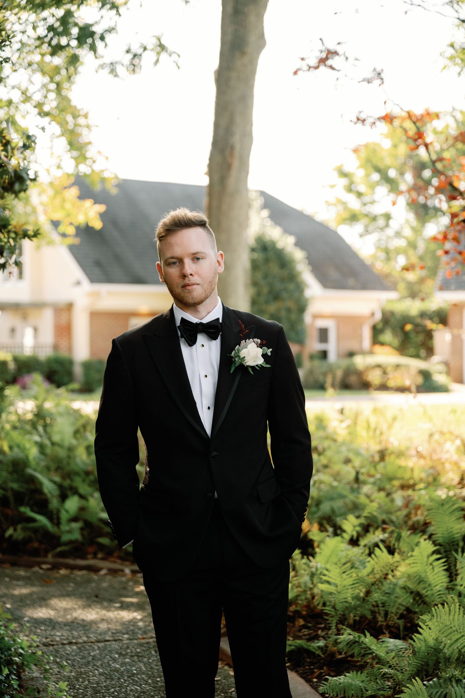 Groom in a black tuxedo with boutonniere poses in the gardens at Church Point Manor in Virginia Beach during his wedding day.