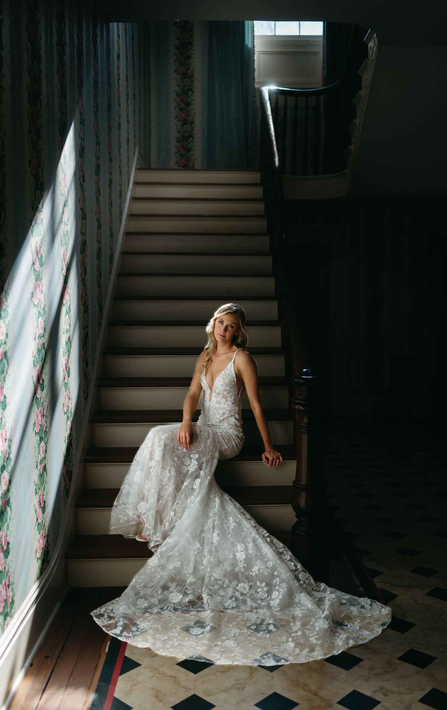 Bride in a lace mermaid gown with a deep V-neckline poses on a grand staircase in dramatic natural light during her Lee Hall wedding in VA