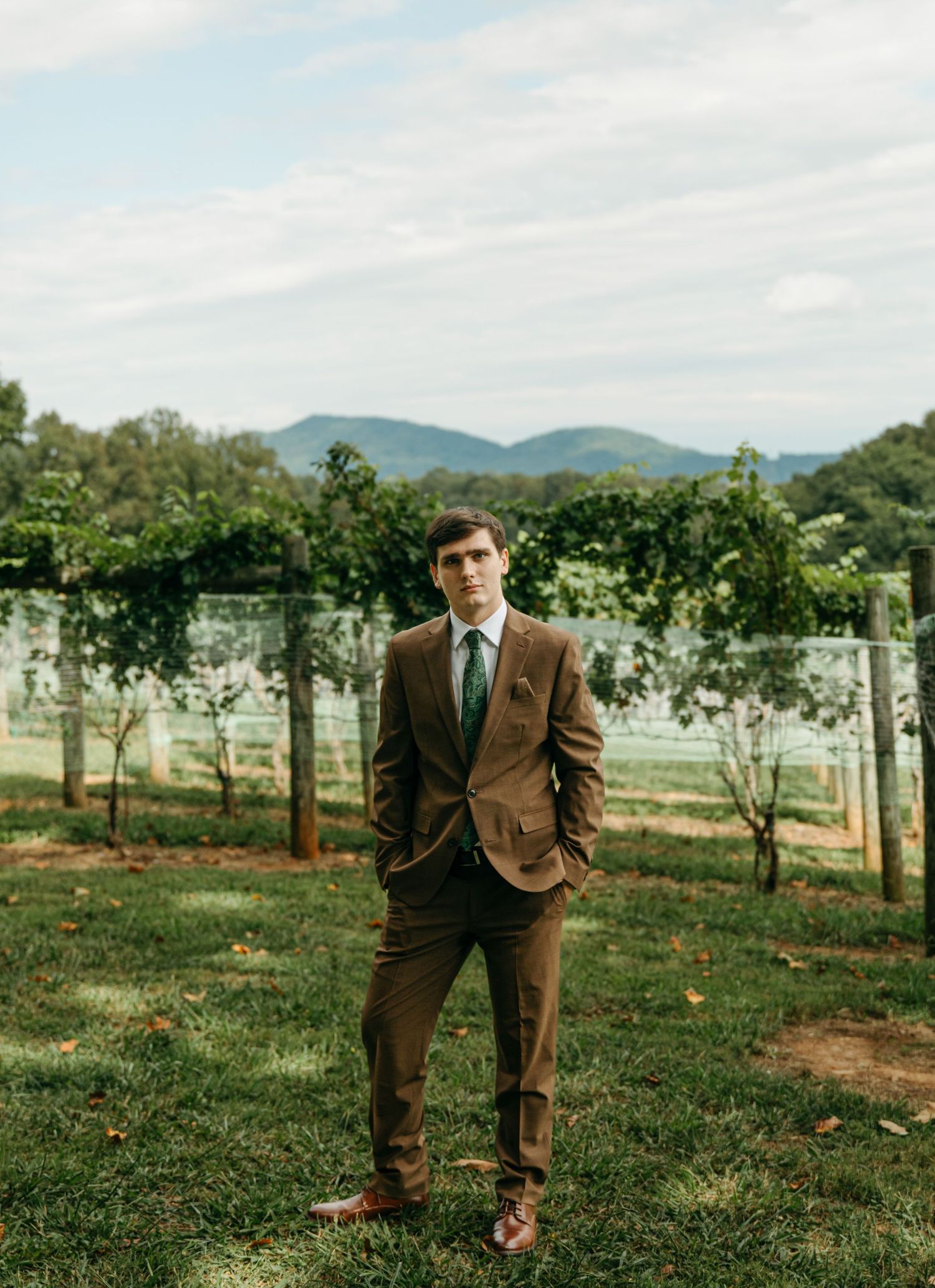 Groom in a brown suit stands confidently in the vineyard with a scenic mountain backdrop on his wedding day at Lovingston Winery in Virginia