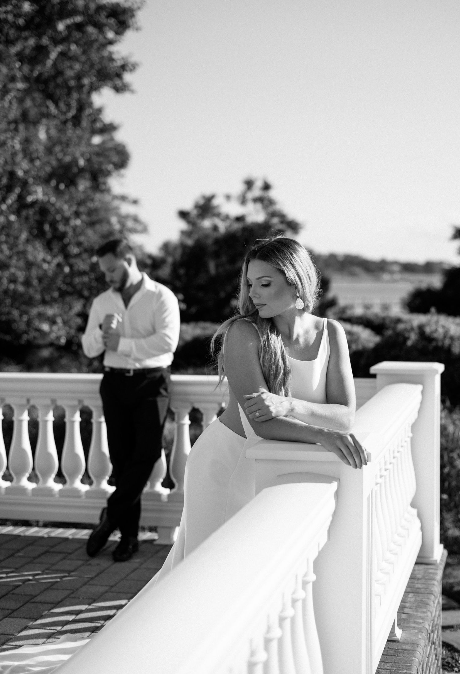 Bride in white gown leans on a balustrade with groom behind her in a black and white wedding portrait at a Virginia estate wedding.