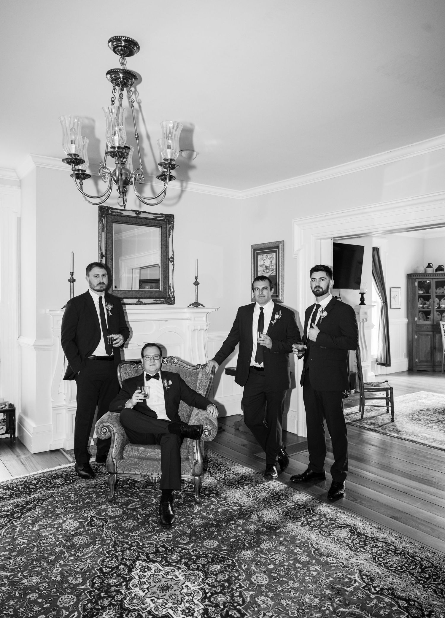 Groomsmen in black tuxedos pose in elegant room in black and white photo.