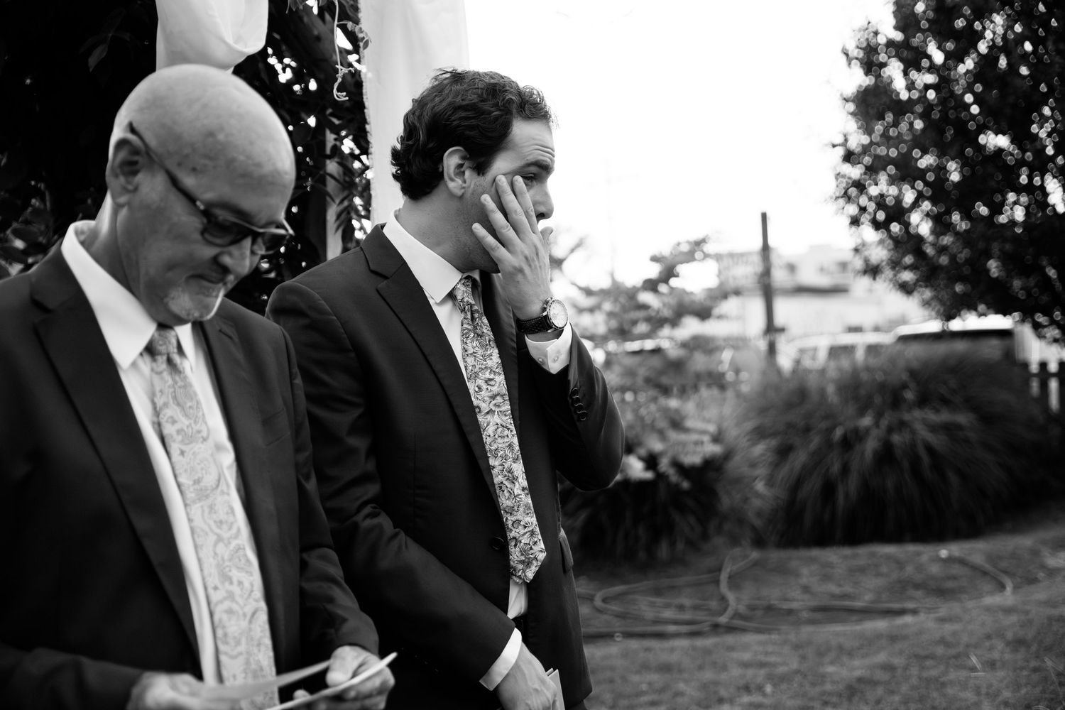 Black and white photo of an emotional groom wiping tears during an outdoor wedding ceremony.