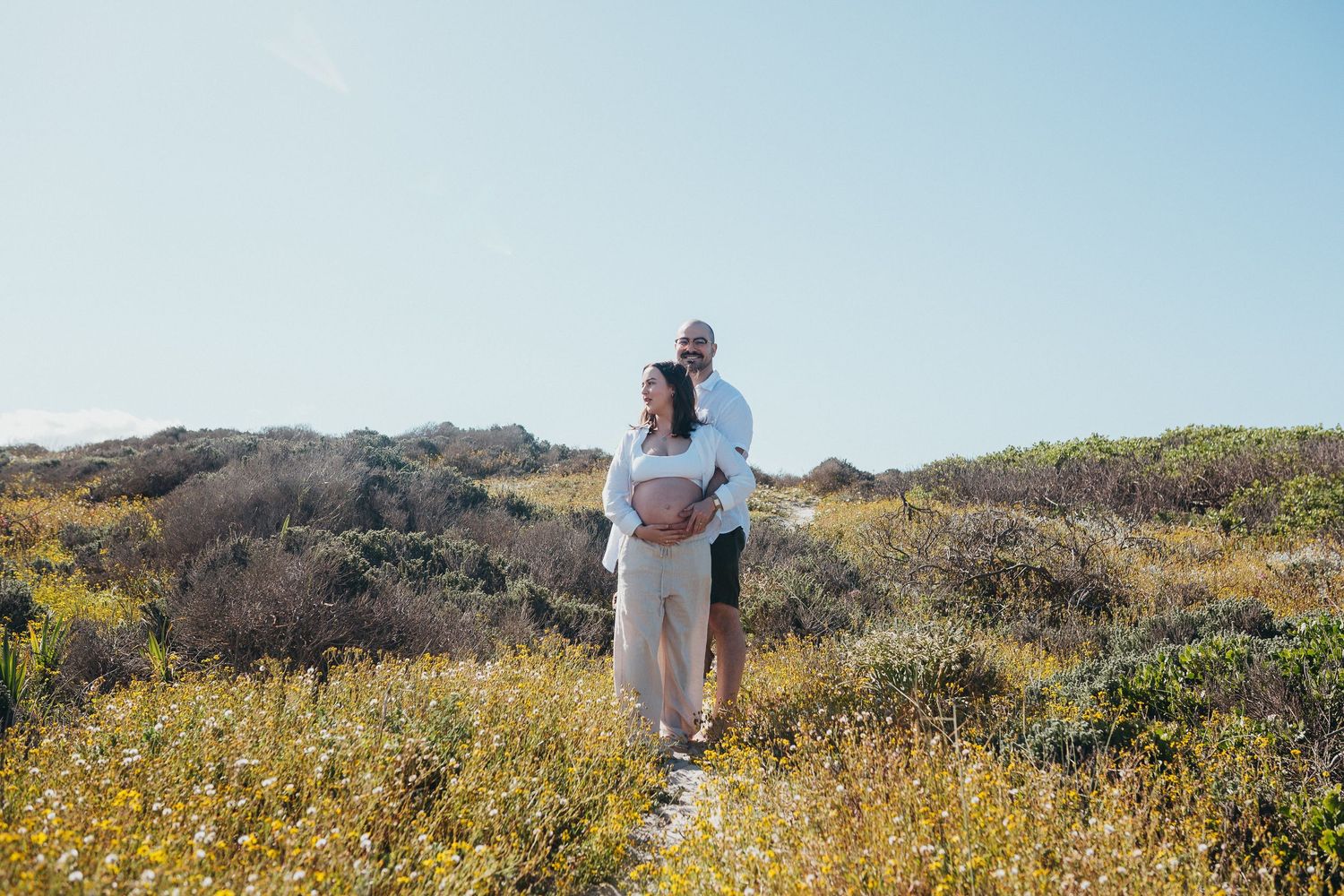 Couple embrace in the wild flowers in their beach maternity shoot