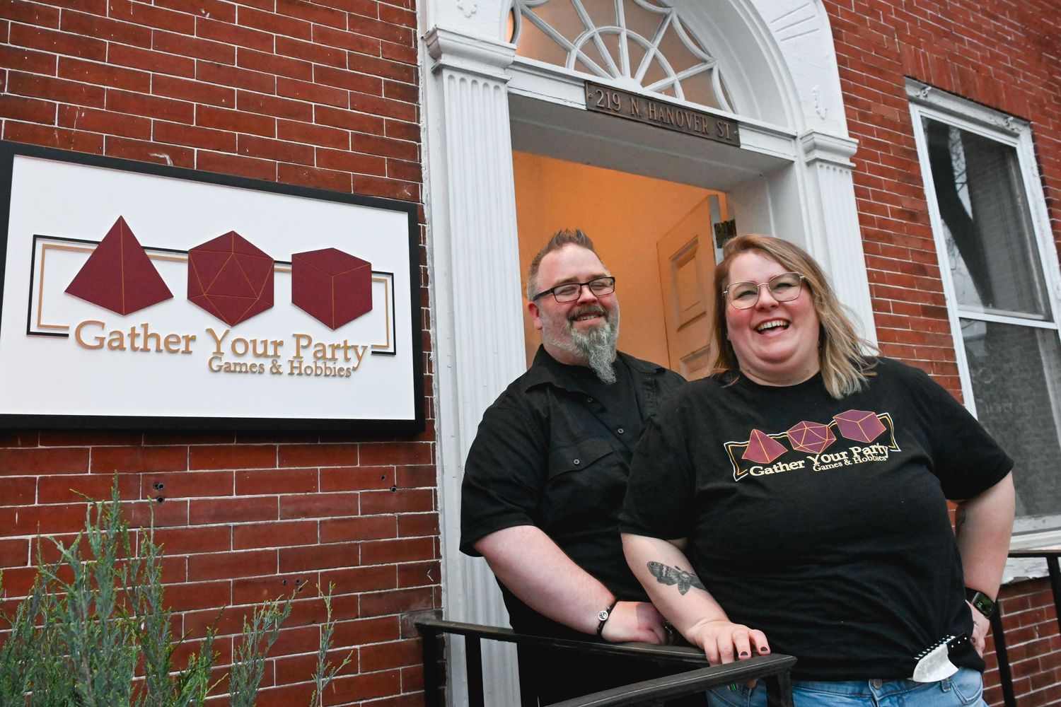 Couple smiling outside Gather Your Party Games and Hobbies store entrance with brick building and logo sign.