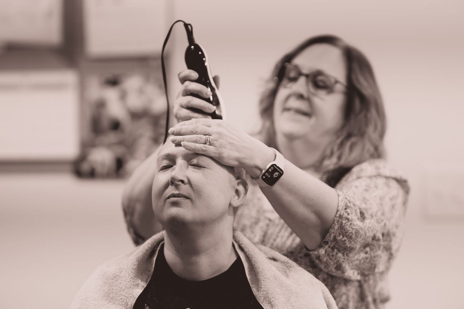 Woman smiling while shaving another persons head with electric clippers in a black and white photo.