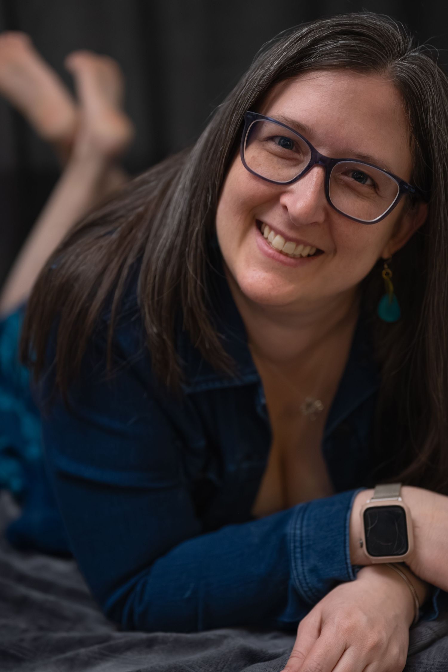 Smiling woman with glasses lying down, wearing denim shirt and smartwatch, with teal earrings.