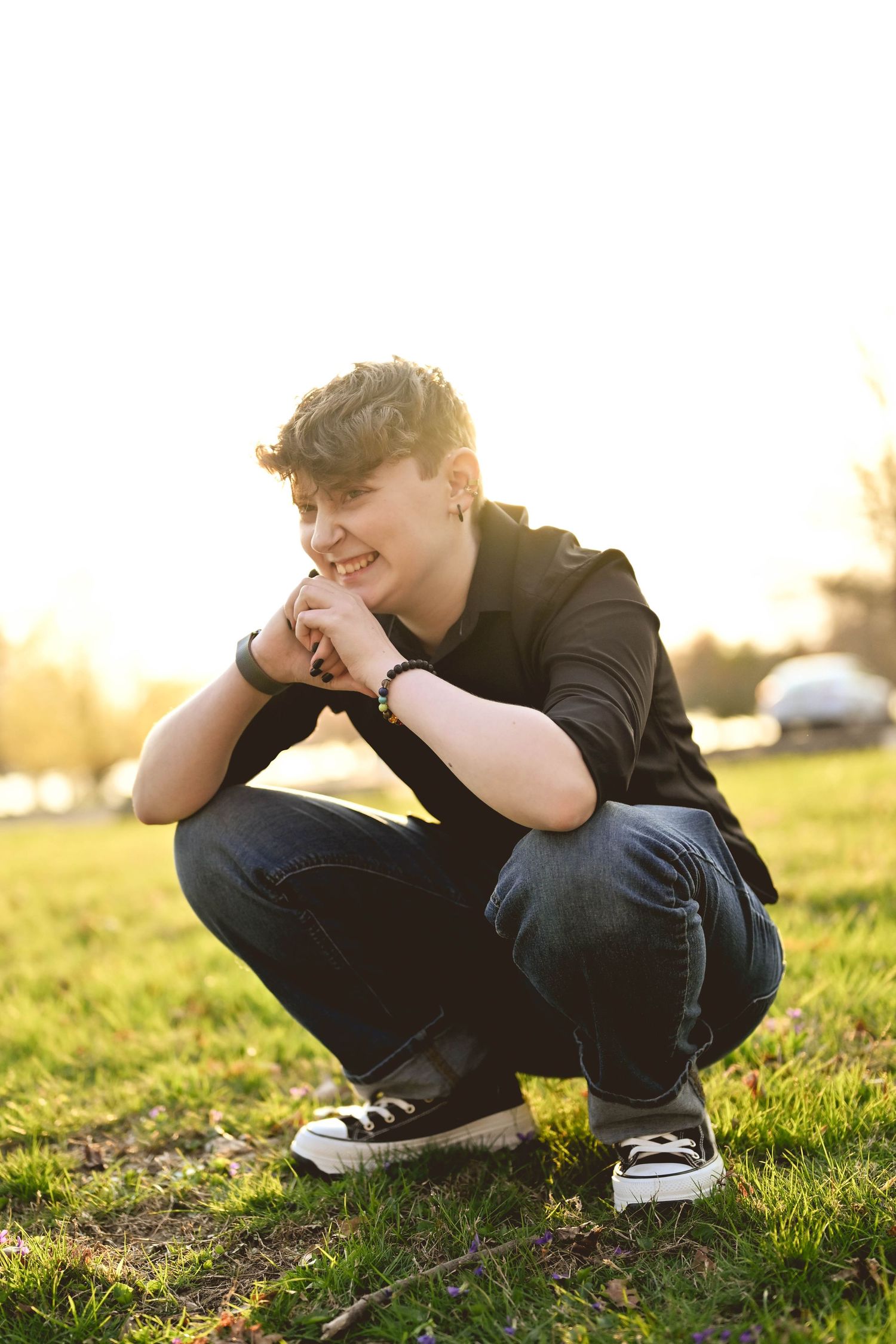 Person in black hoodie and jeans crouches on grassy field during golden sunset hour while looking down thoughtfully.