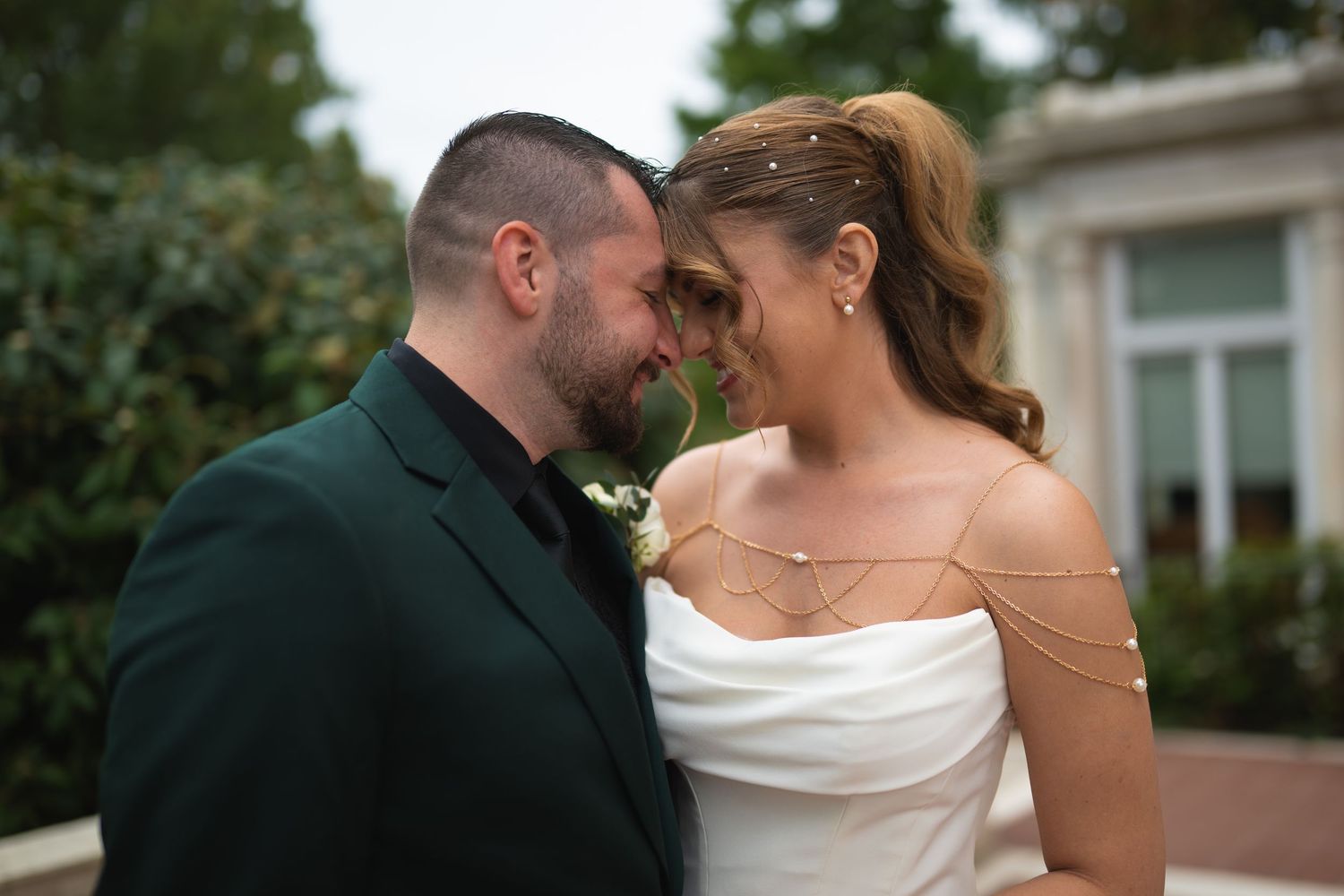 Bride in a white strapless wedding dress with draped body chains touches foreheads with groom in a dark green suit outdoors.