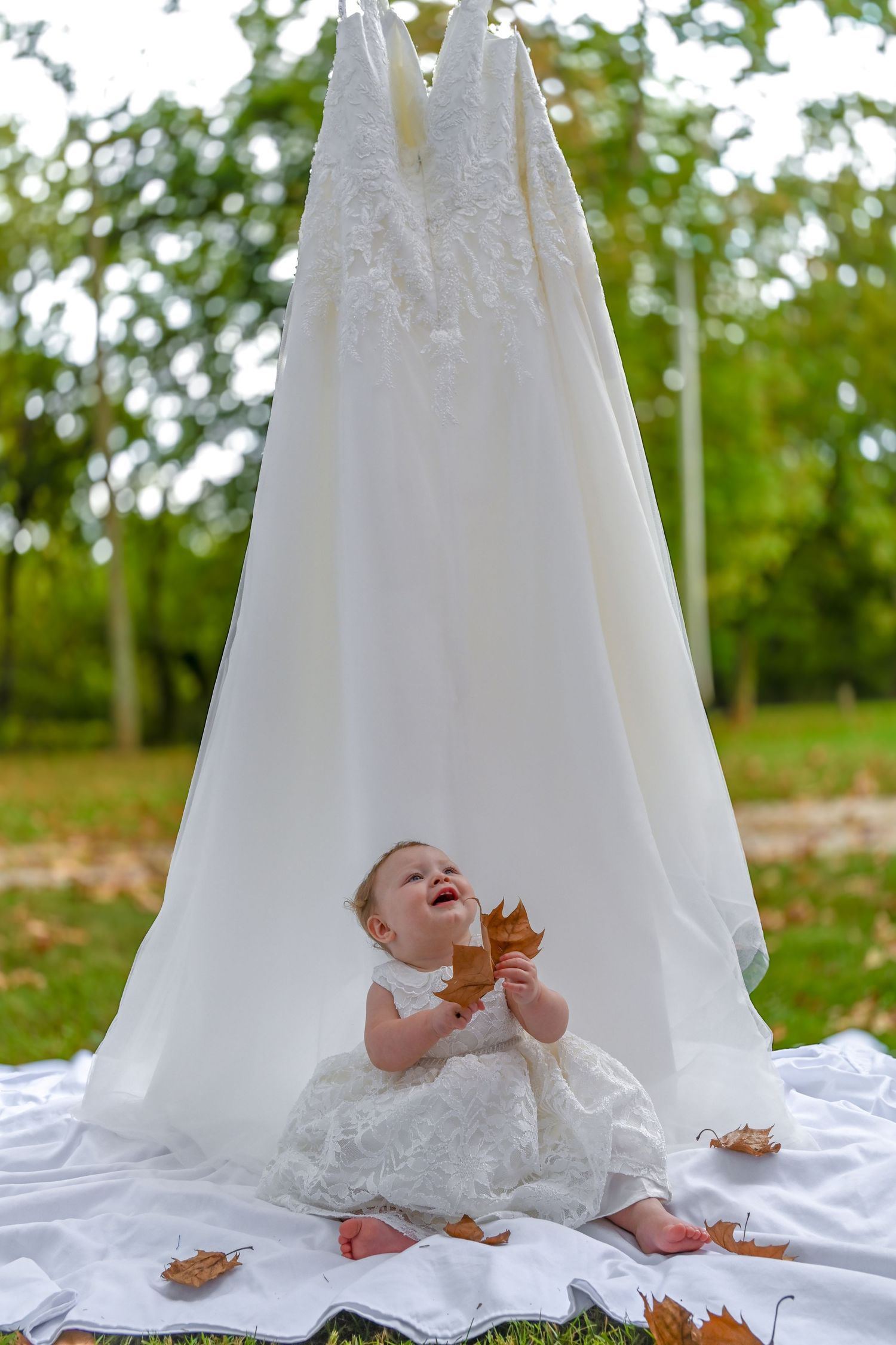 Baby girl in a white lace dress sits on a blanket holding autumn leaves beneath a hanging wedding gown in a sunny park.