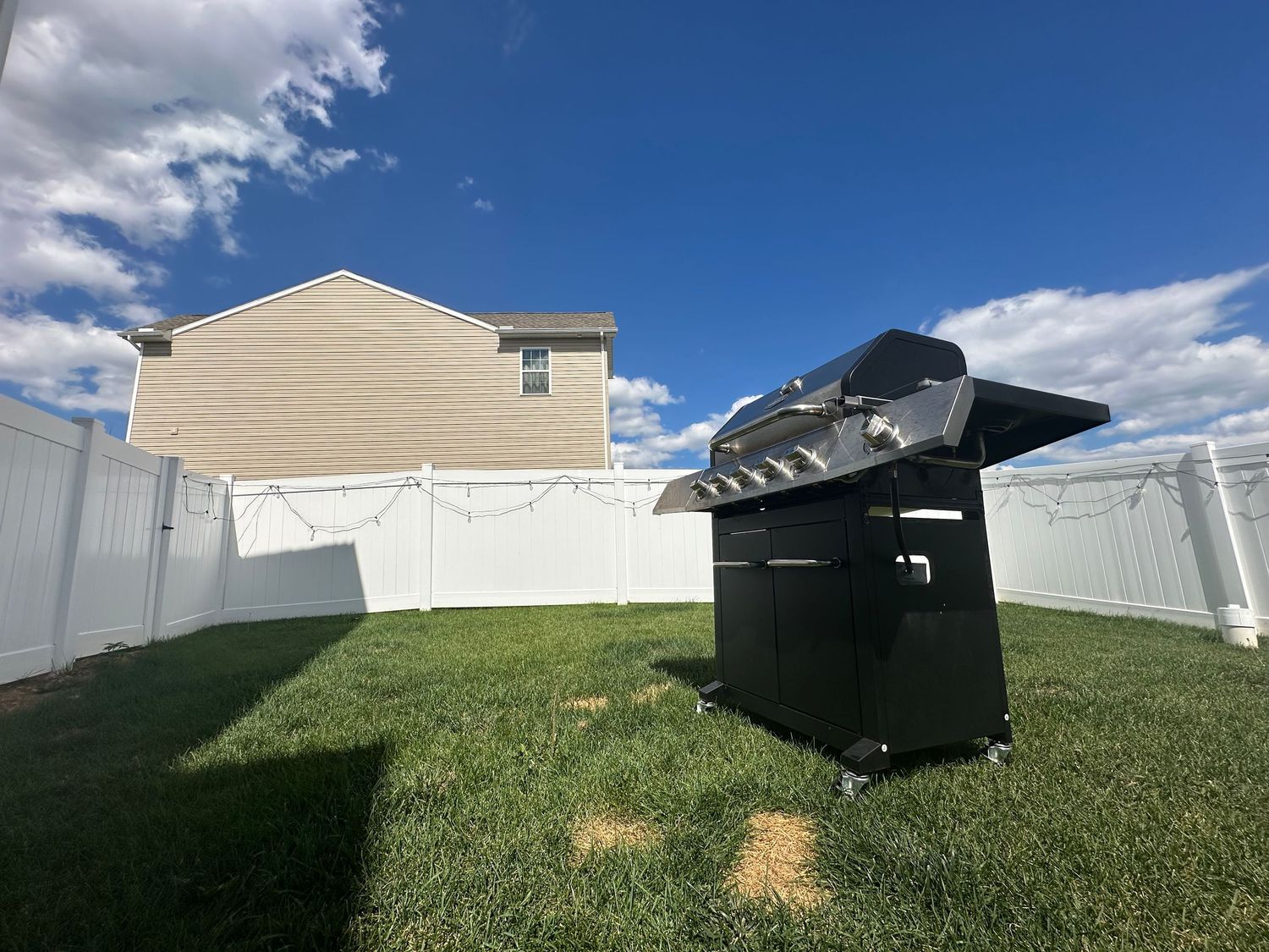 Black gas grill on green lawn in fenced backyard with white vinyl fence and neighboring house under blue sky.