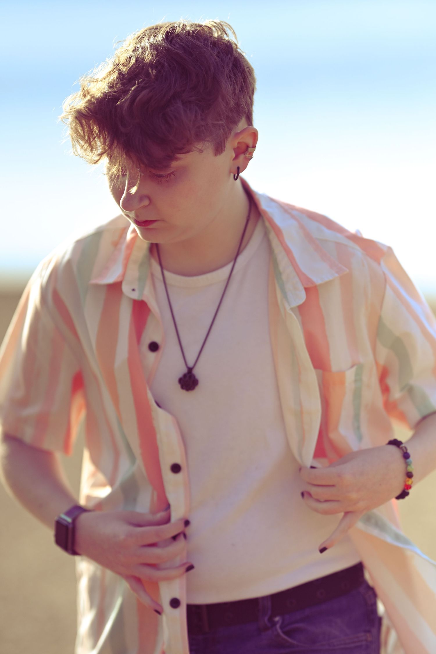 Young person in sunlit outdoor setting wearing white t-shirt, pink button-up shirt and necklace with soft sepia tones.