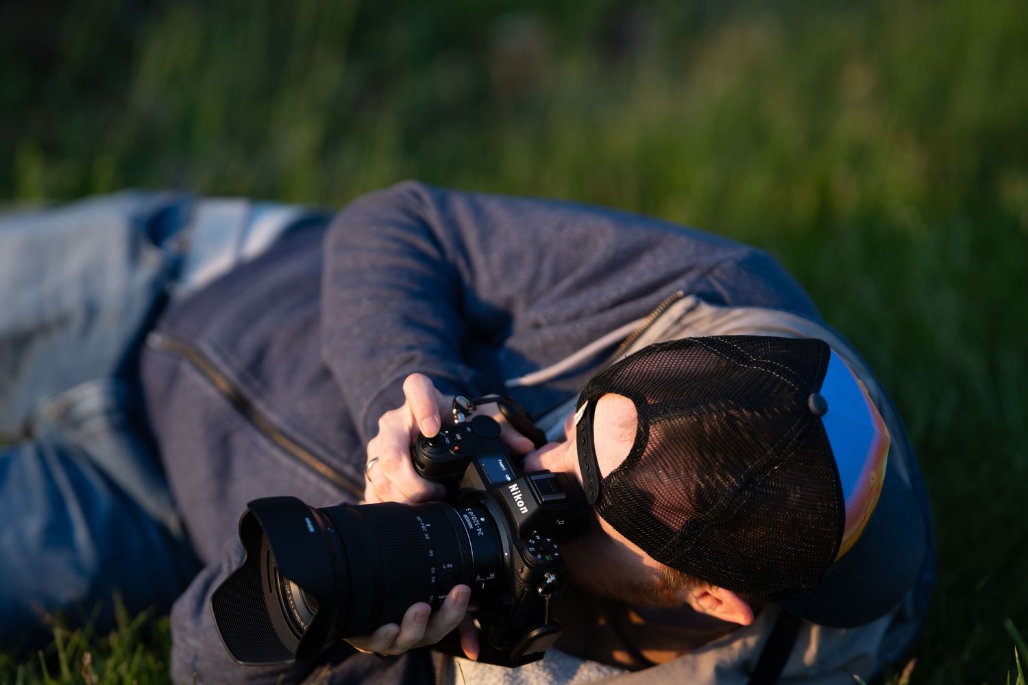 Photographer lying on grass in golden hour light, capturing close-up shots with a DSLR camera and large lens.