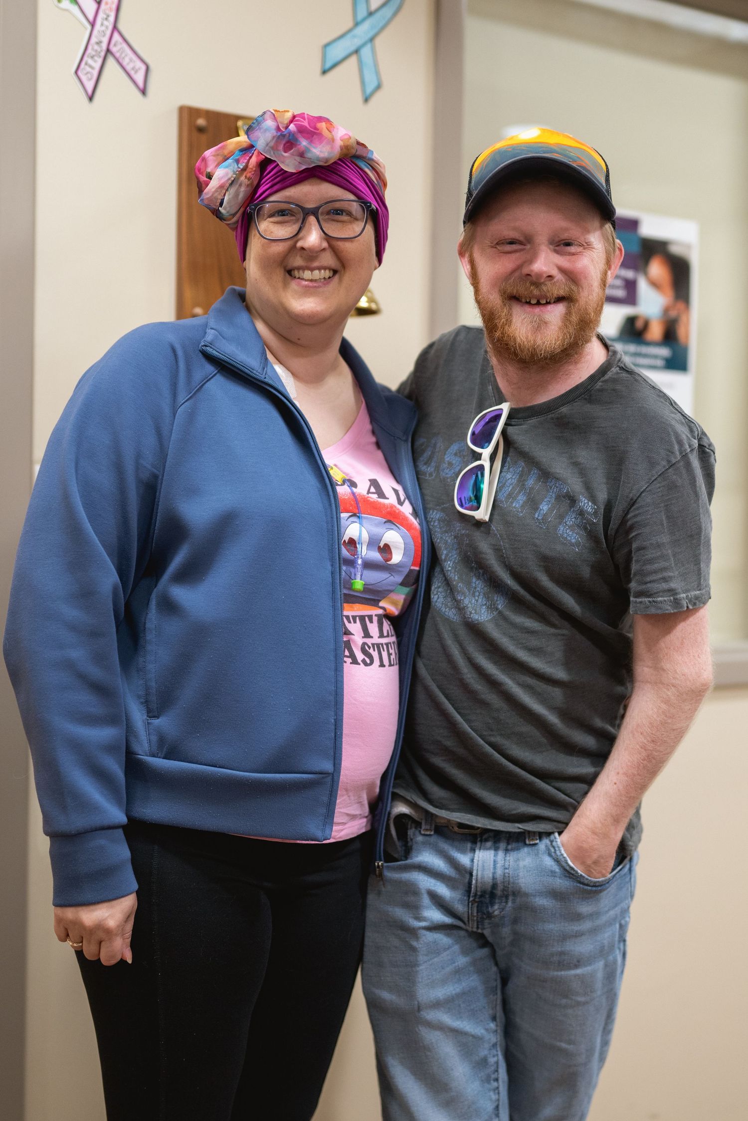 A smiling couple poses together indoors, woman wearing colorful headscarf and blue jacket, man in gray t-shirt and cap.