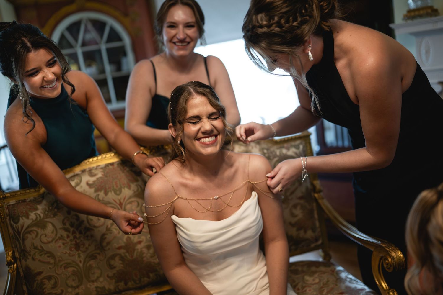 Bridesmaids help a smiling bride with her necklace while getting ready in an elegant room.