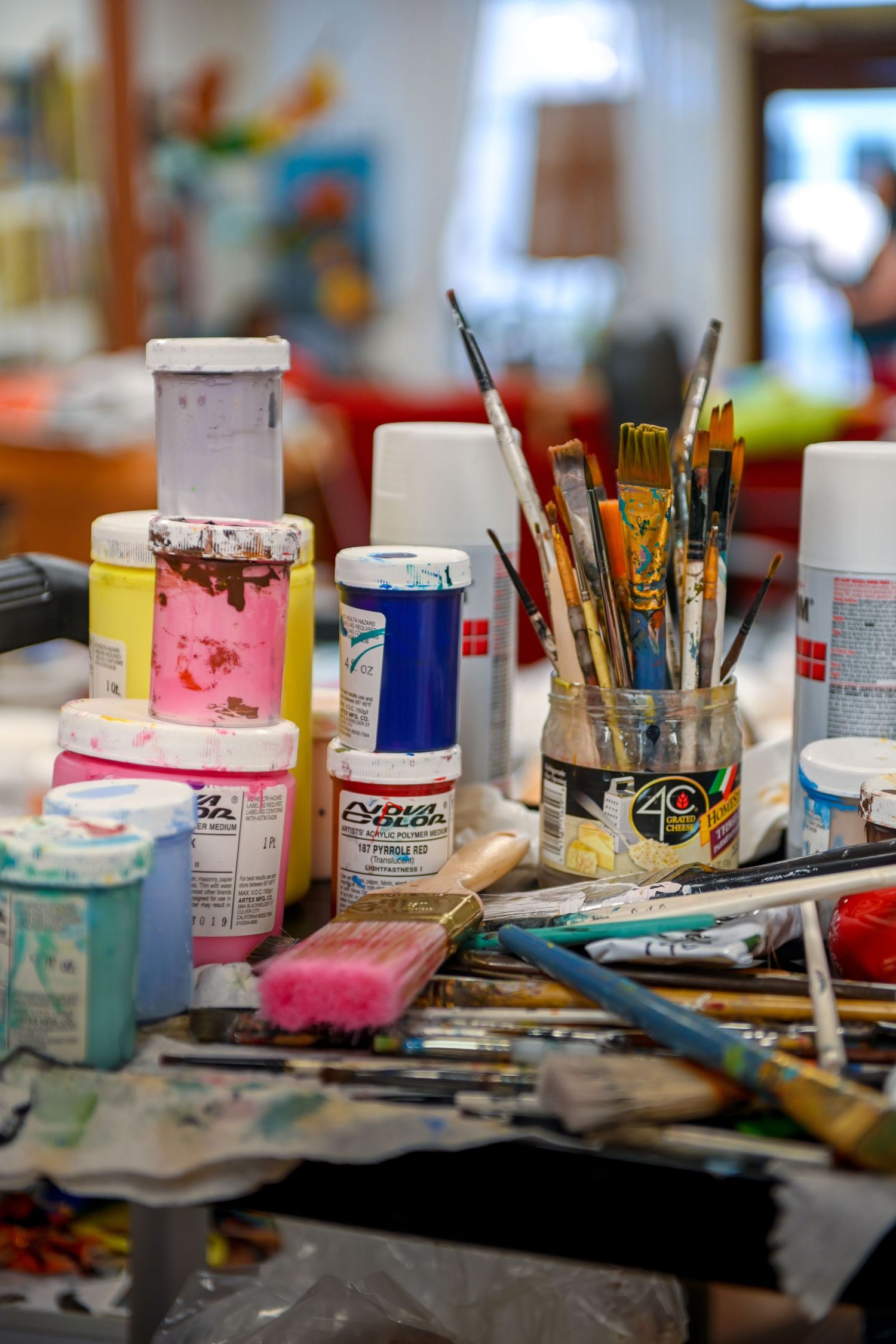 A messy artist's workspace with paint jars, brushes, and art supplies scattered on a surface in natural lighting.
