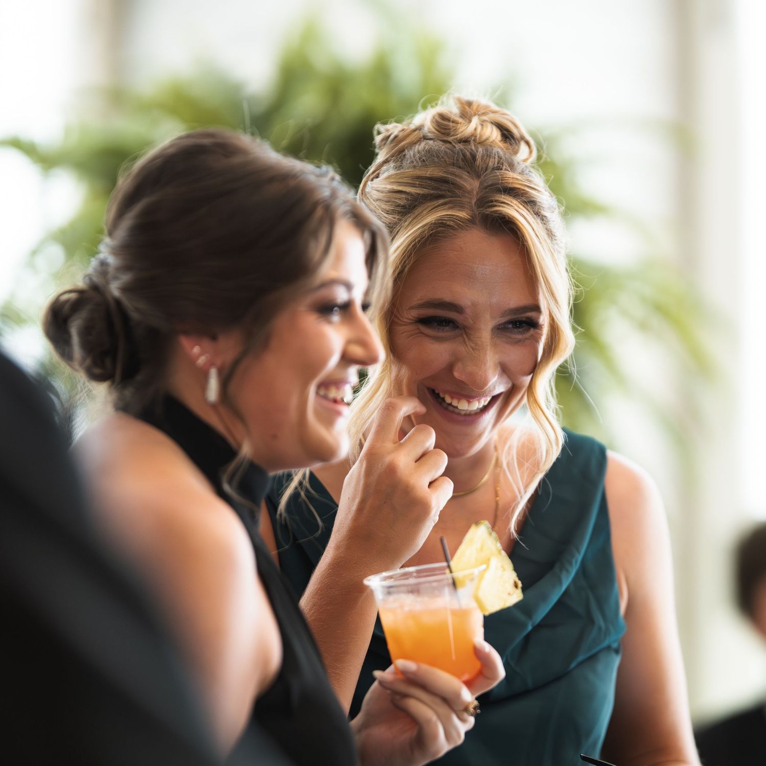 Two women laughing and enjoying drinks at a wedding reception, wearing elegant green dresses.