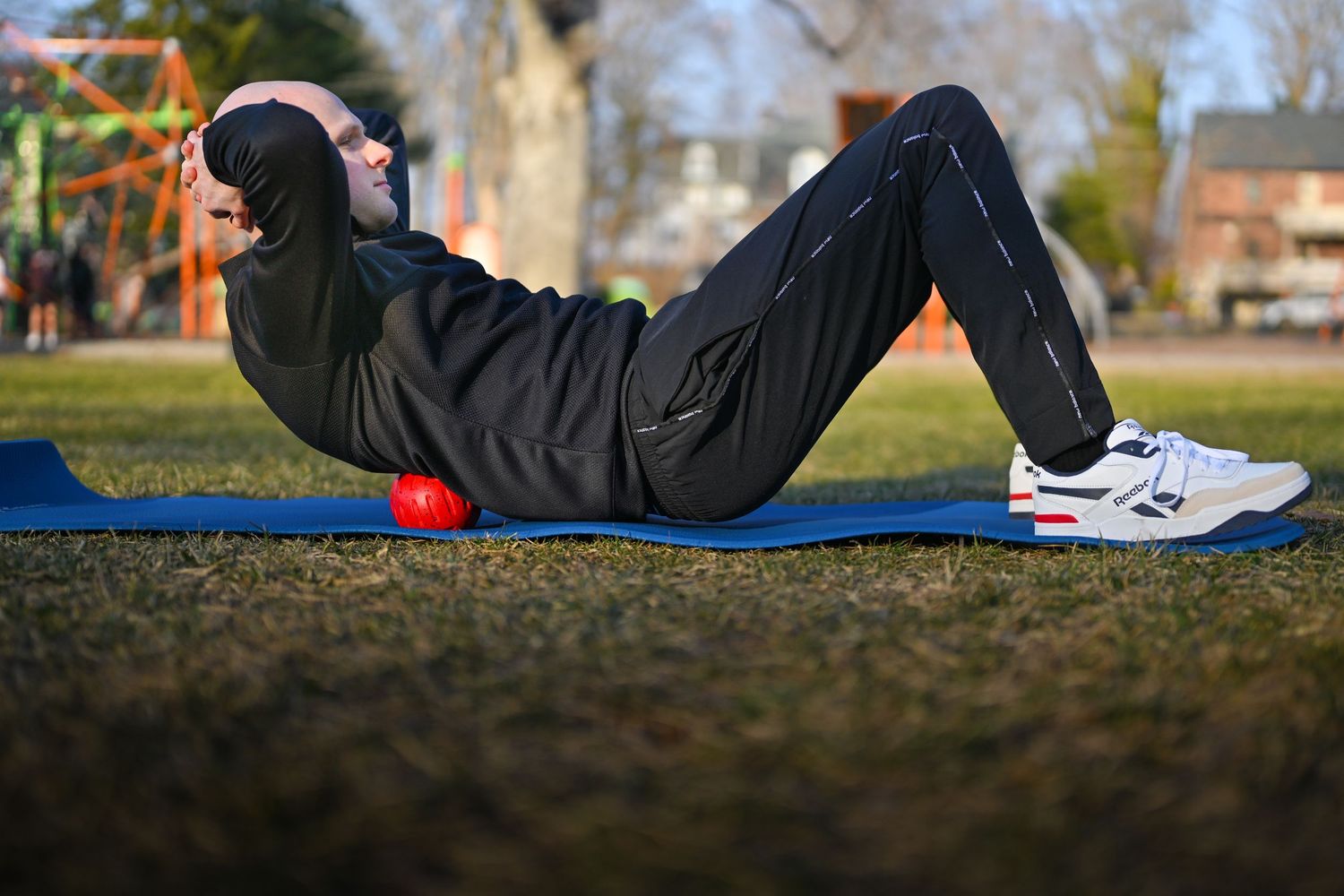 Someone doing core exercises on a blue yoga mat outdoors while wearing a black hoodie and athletic shoes.