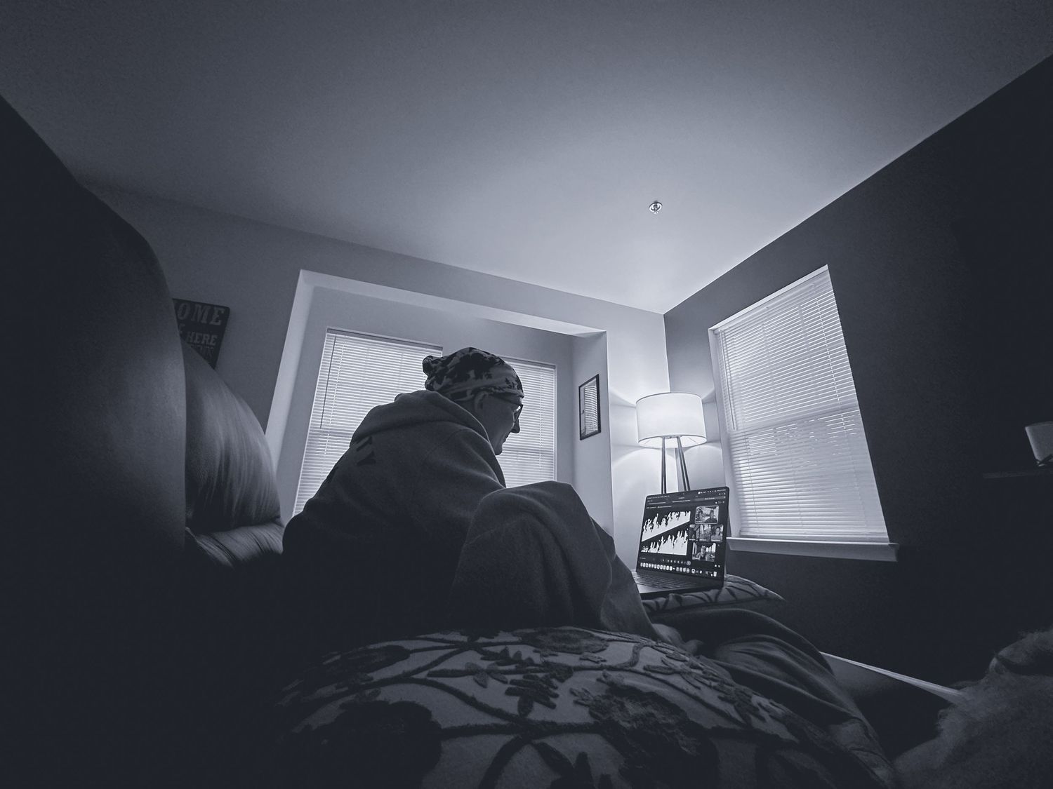 Solitary person sitting on bed in dark room, black and white photo capturing loneliness and contemplation near window.