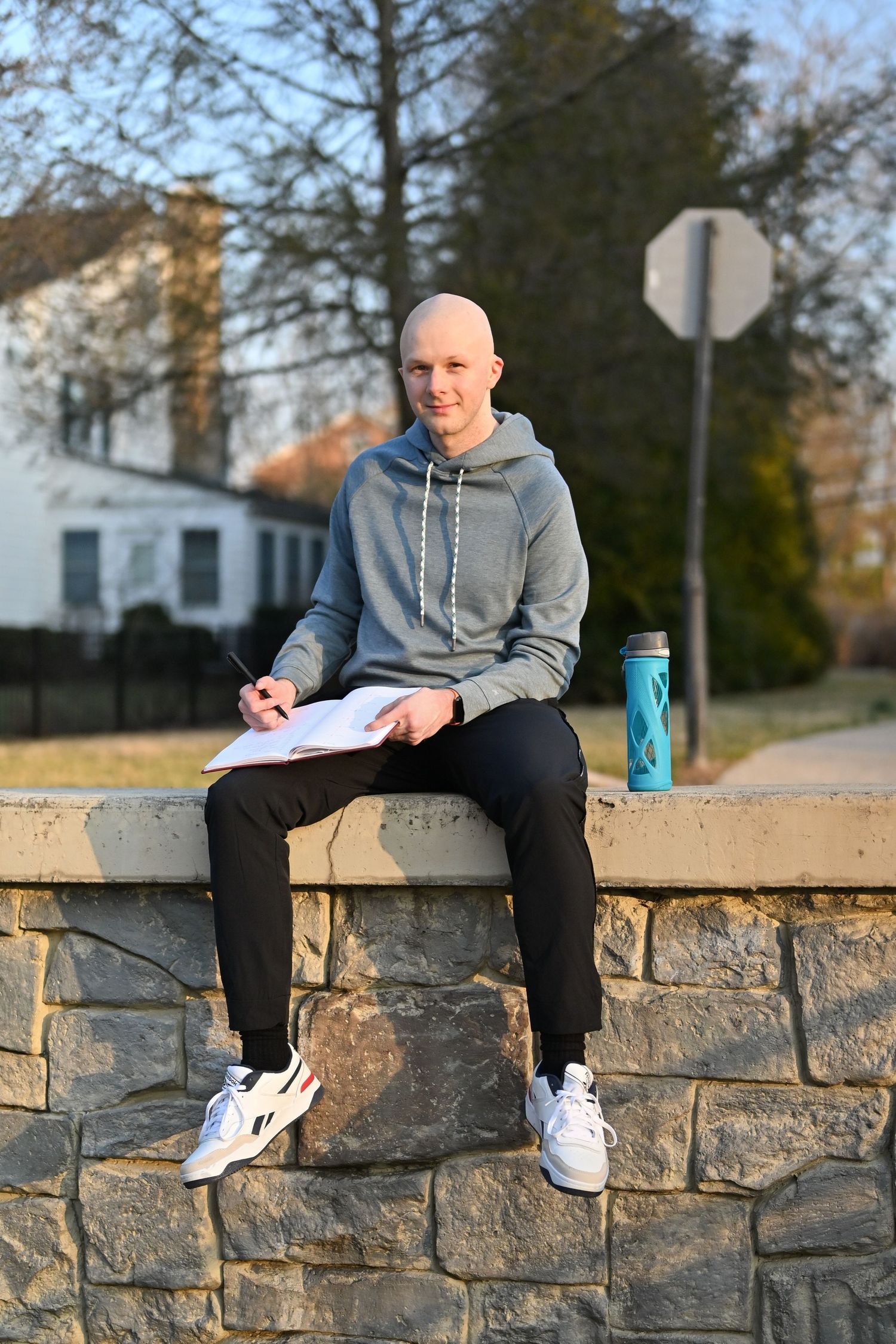 Person in grey hoodie sitting on stone wall with notebook and water bottle.