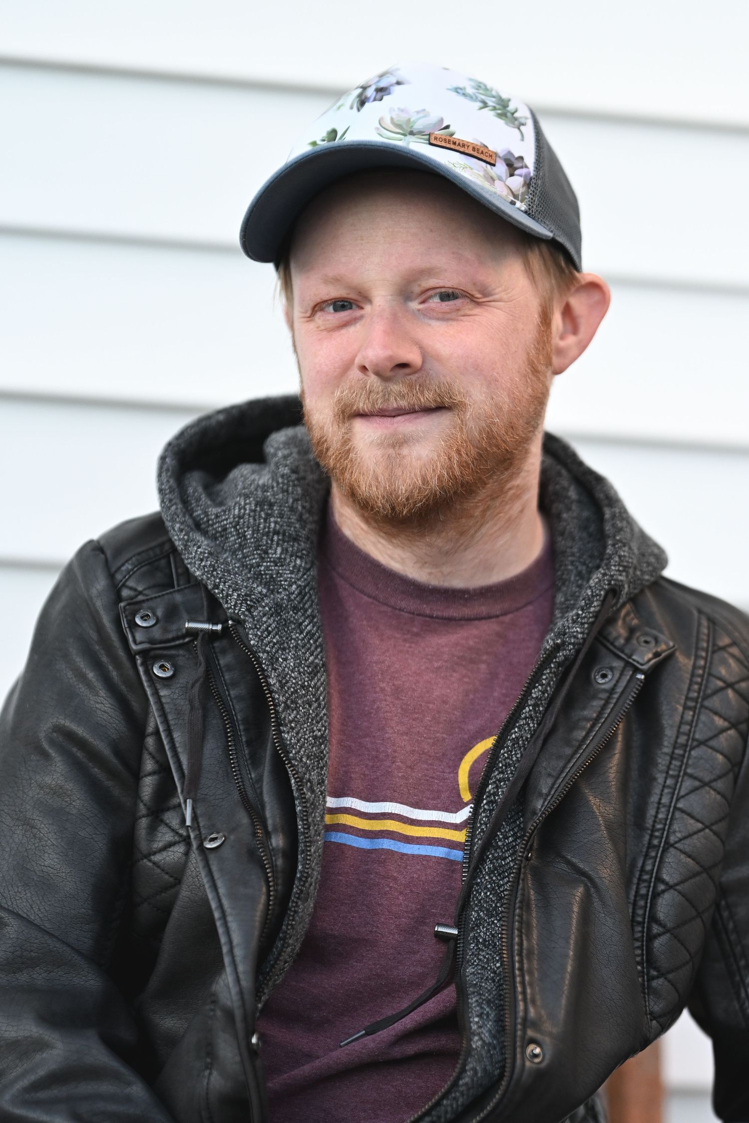 Person wearing a leather jacket and baseball cap with a red beard smiles against white siding outdoors.