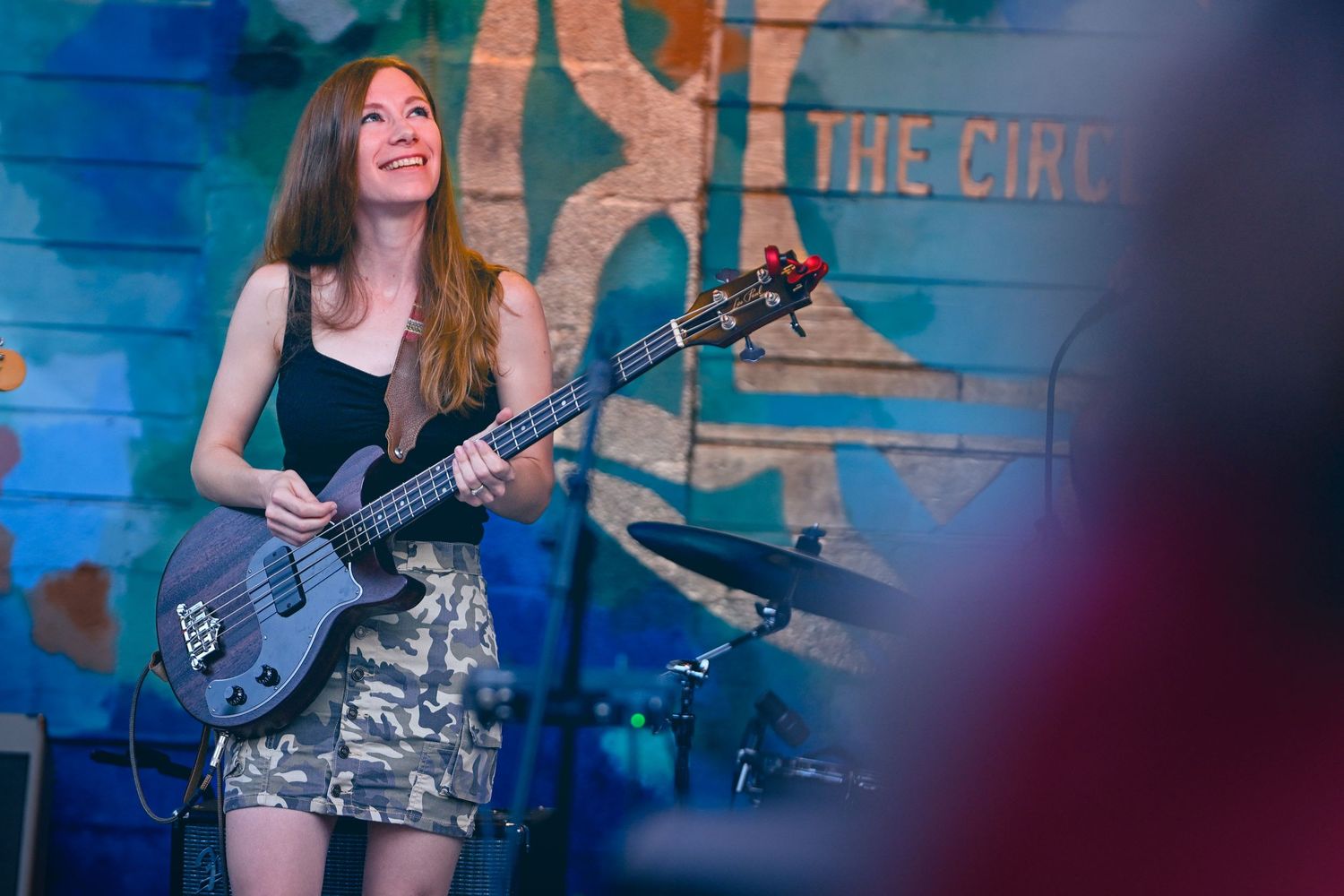 Musician playing electric bass guitar performs on stage with colorful blue backdrop during live concert performance.