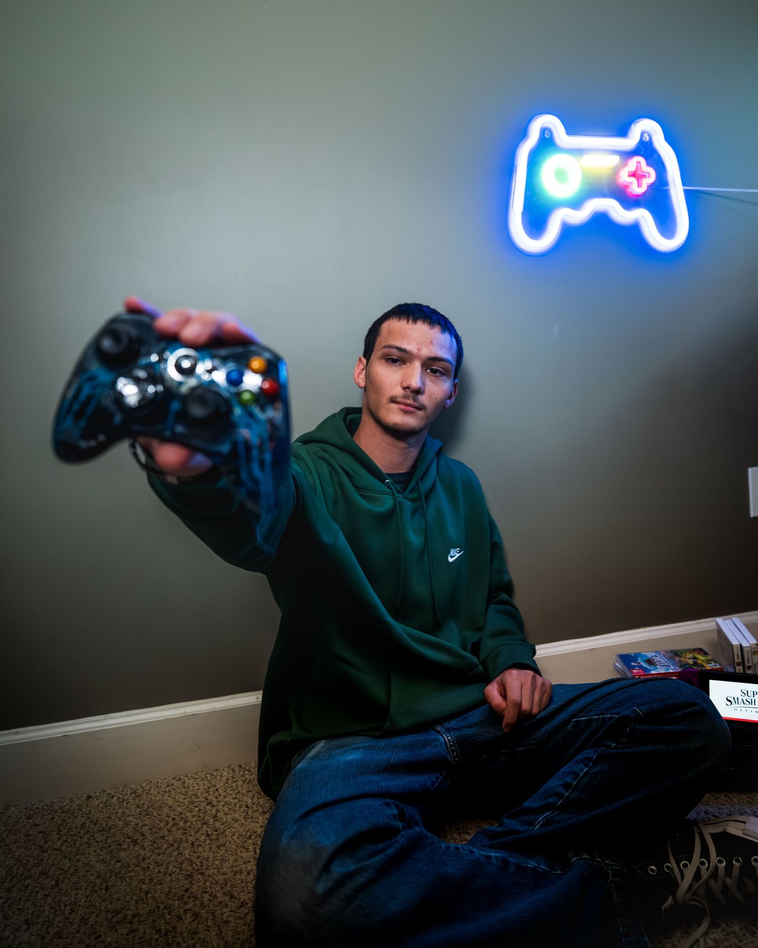 Young man in a green hoodie sits on the floor holding out a game controller toward the camera beside a glowing neon gamepad sign.