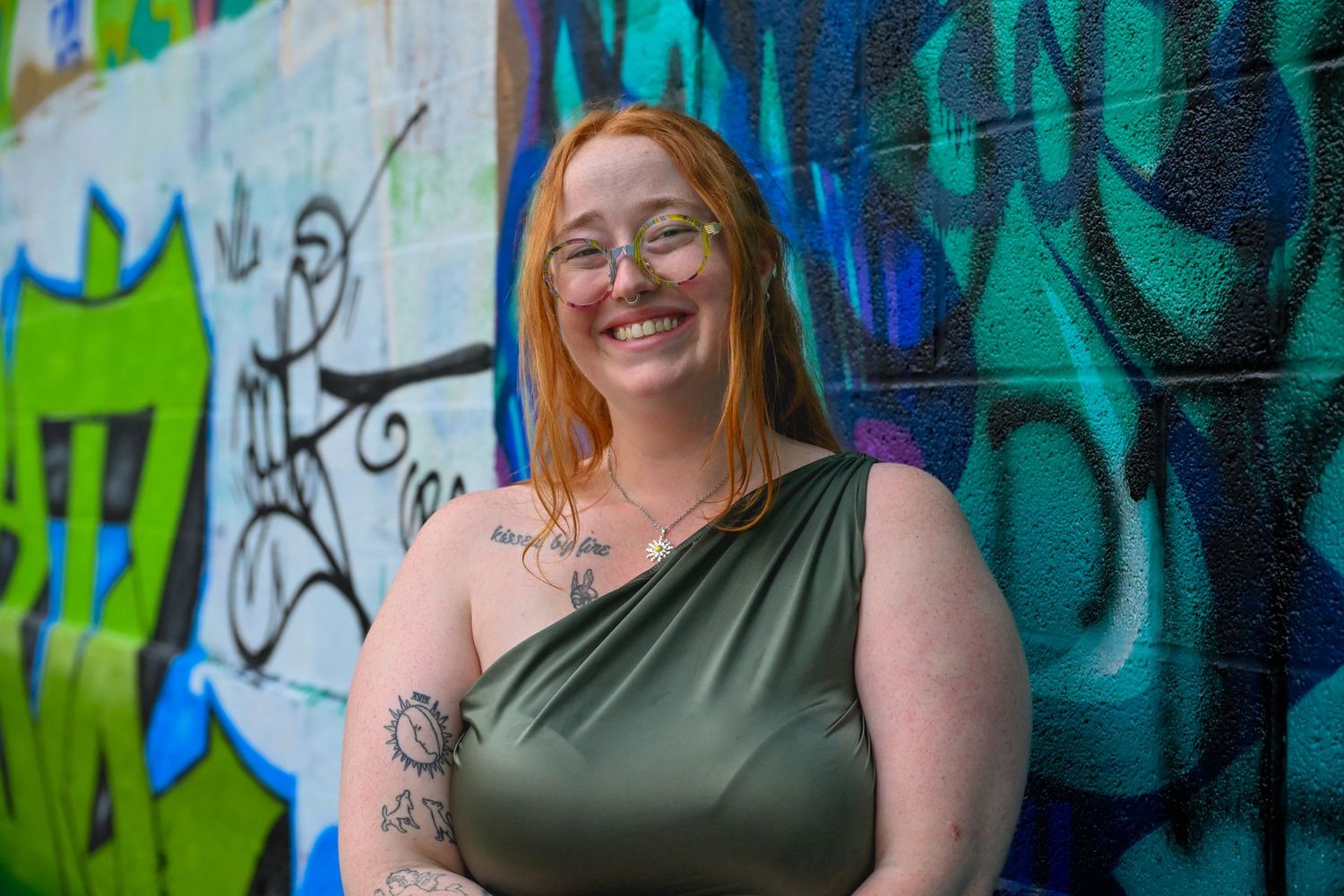 A smiling person with red hair and tattoos poses against a vibrant graffiti wall wearing a one-shoulder green top.