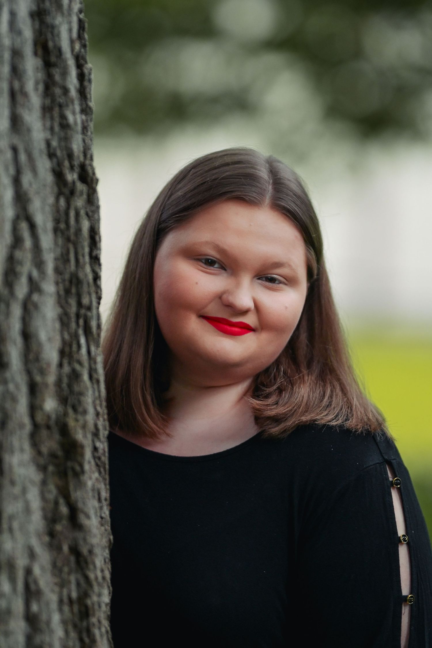Portrait of a person smiling warmly while standing beside a tree trunk outdoors, wearing a black top and red lipstick.