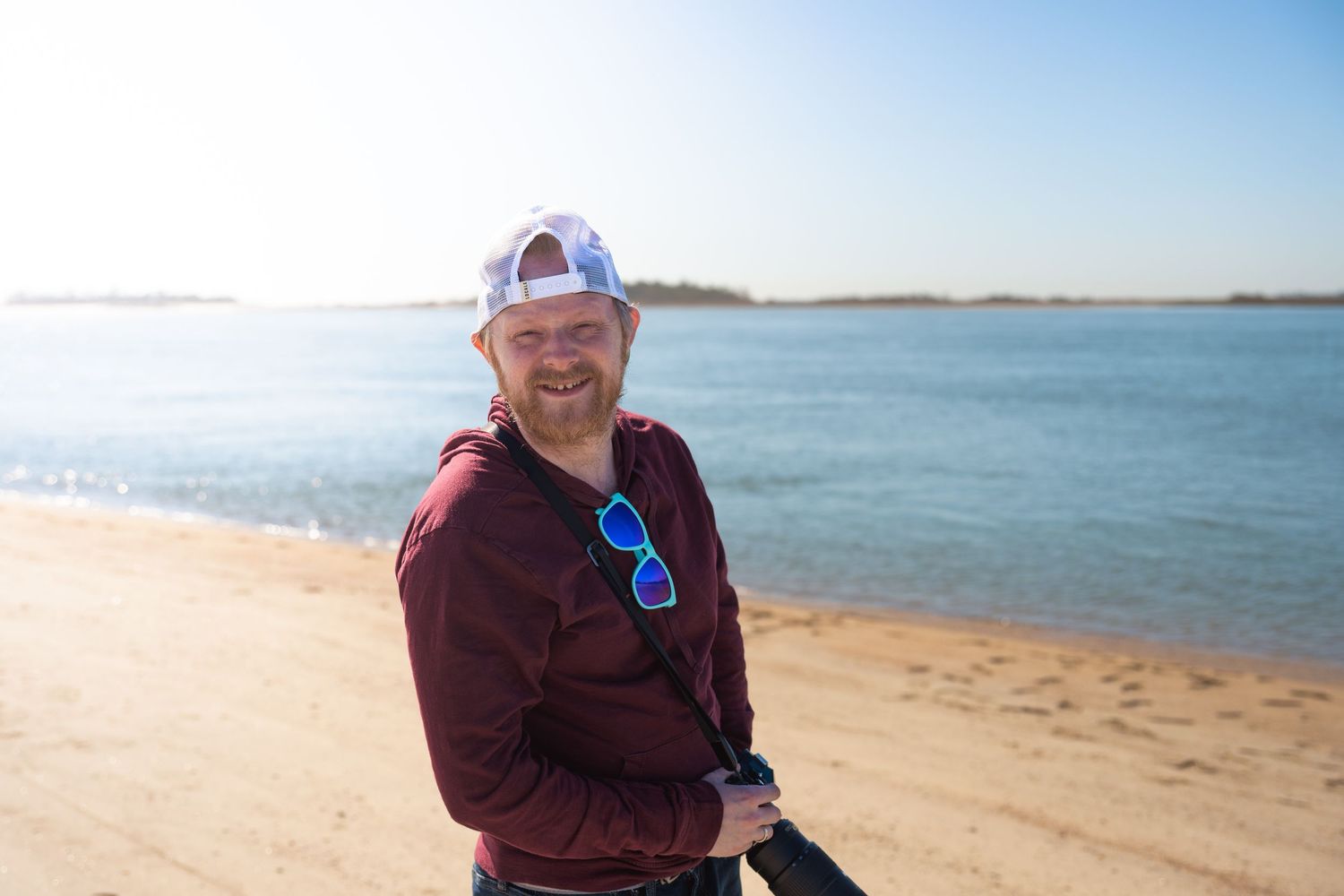 A person walks along a sandy beach on a sunny day with calm blue waters and clear skies in the background.