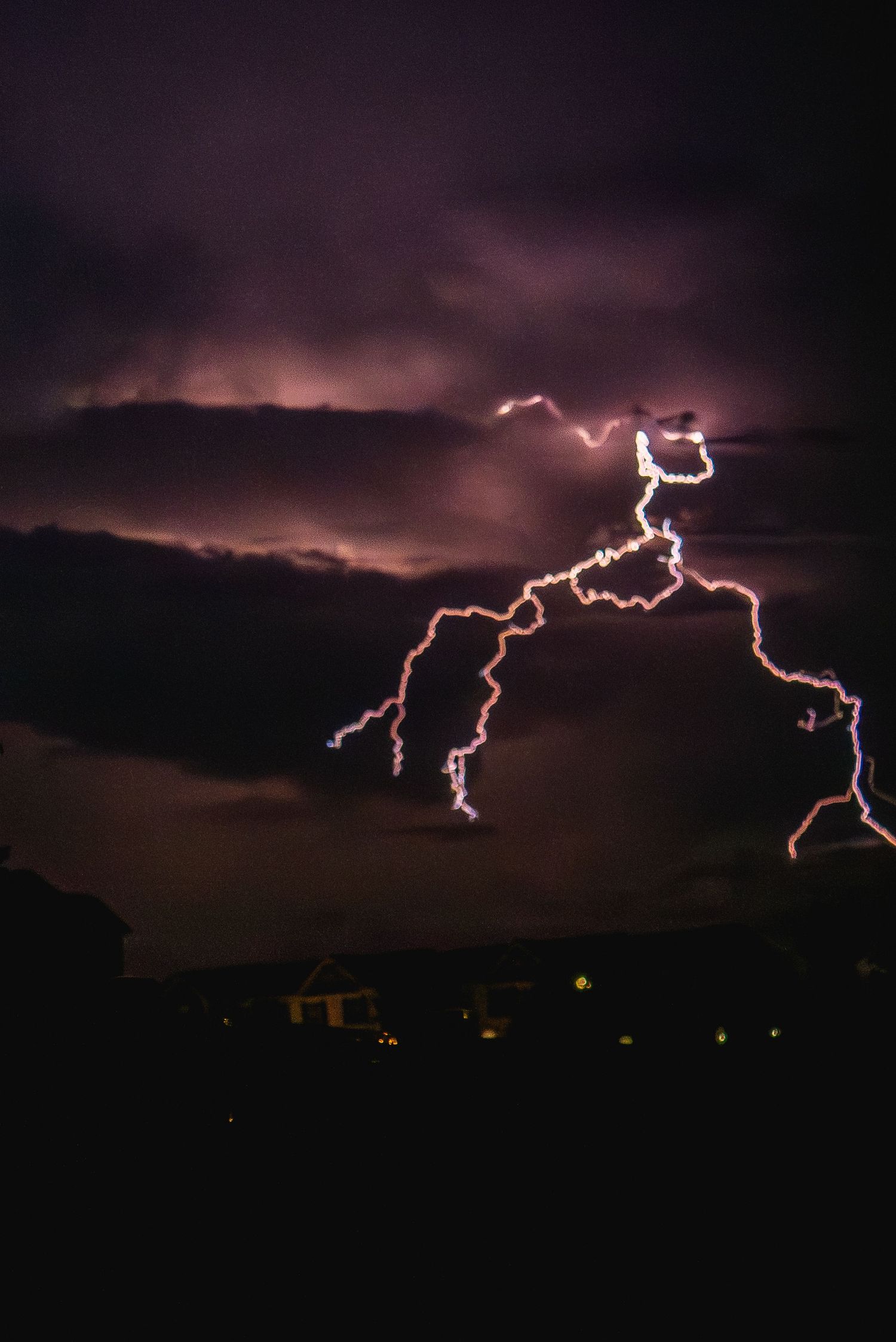 Dramatic lightning bolt branching across a dark purple stormy night sky illuminating clouds above a silhouetted cityscape.