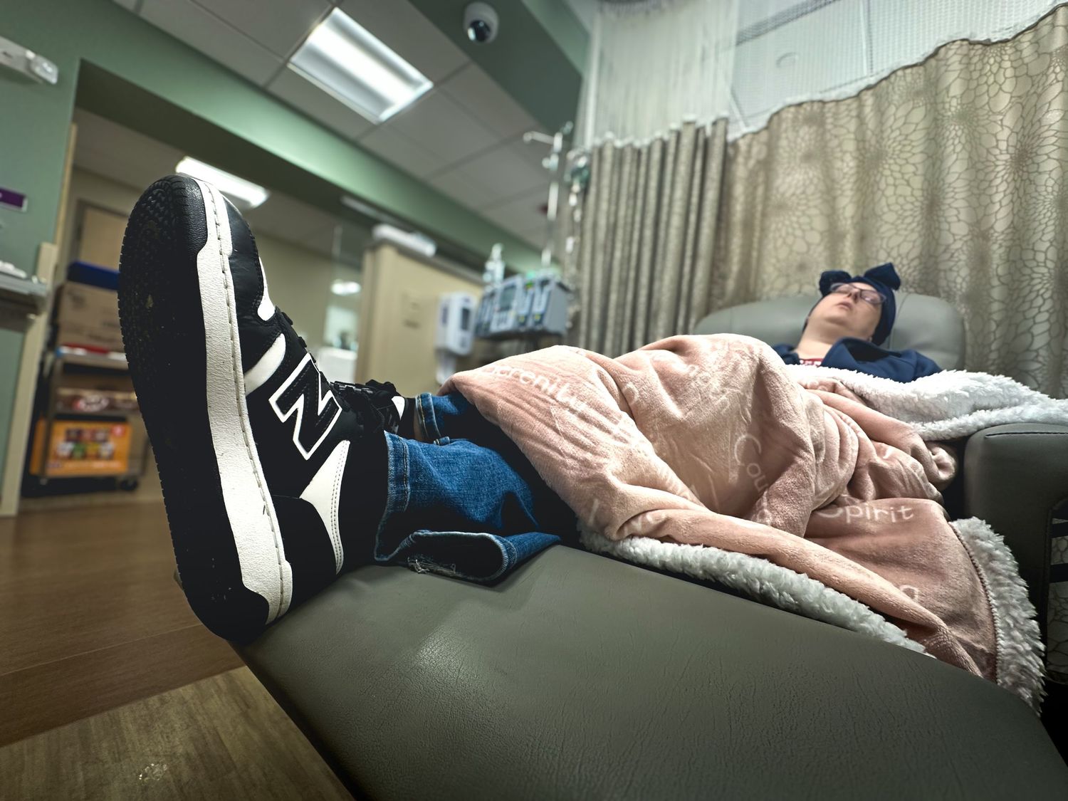 Person resting on a medical chair wearing New Balance sneakers and jeans, covered with a pink blanket in a clinic.
