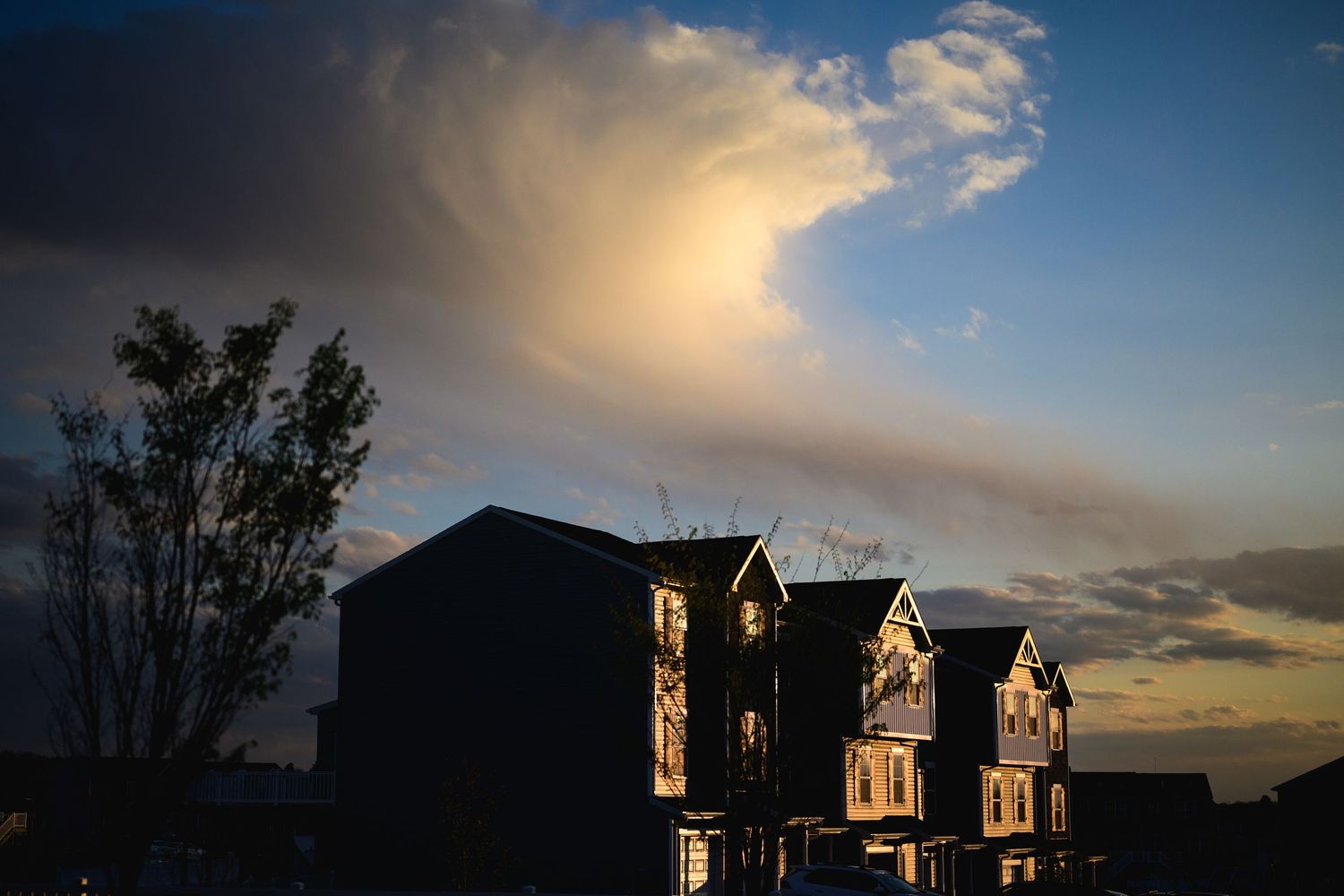 Silhouetted suburban homes at sunset with dramatic storm clouds illuminated by golden light in the evening sky.