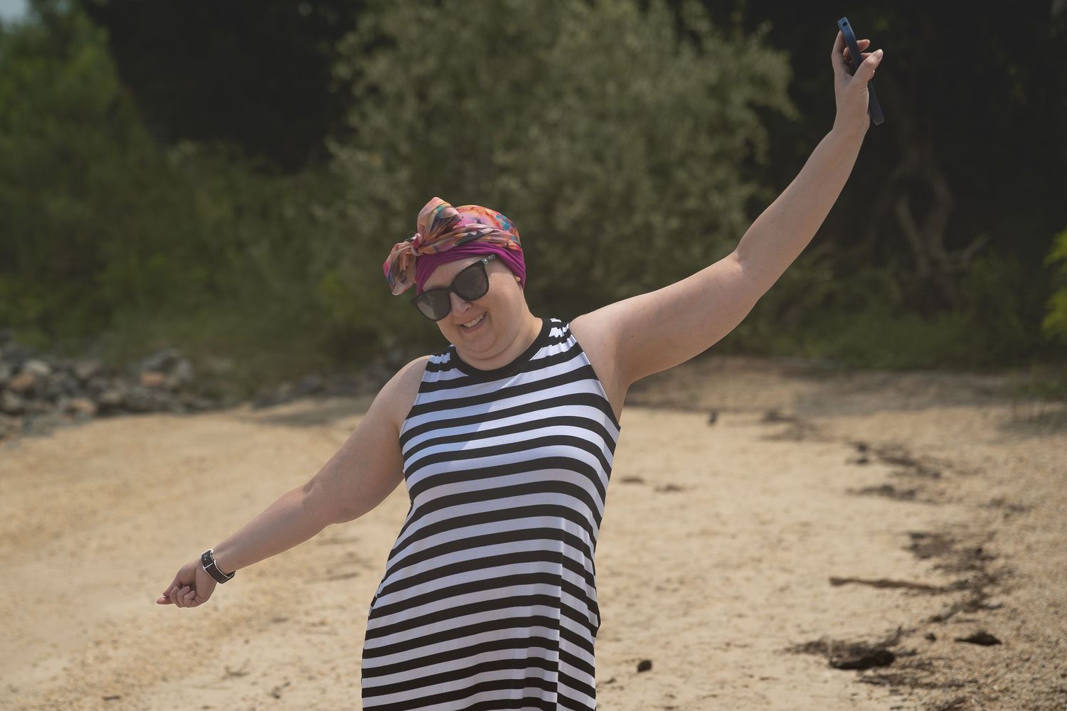 Woman in striped dress and red headband dancing joyfully on a sandy path with arms raised outdoors.