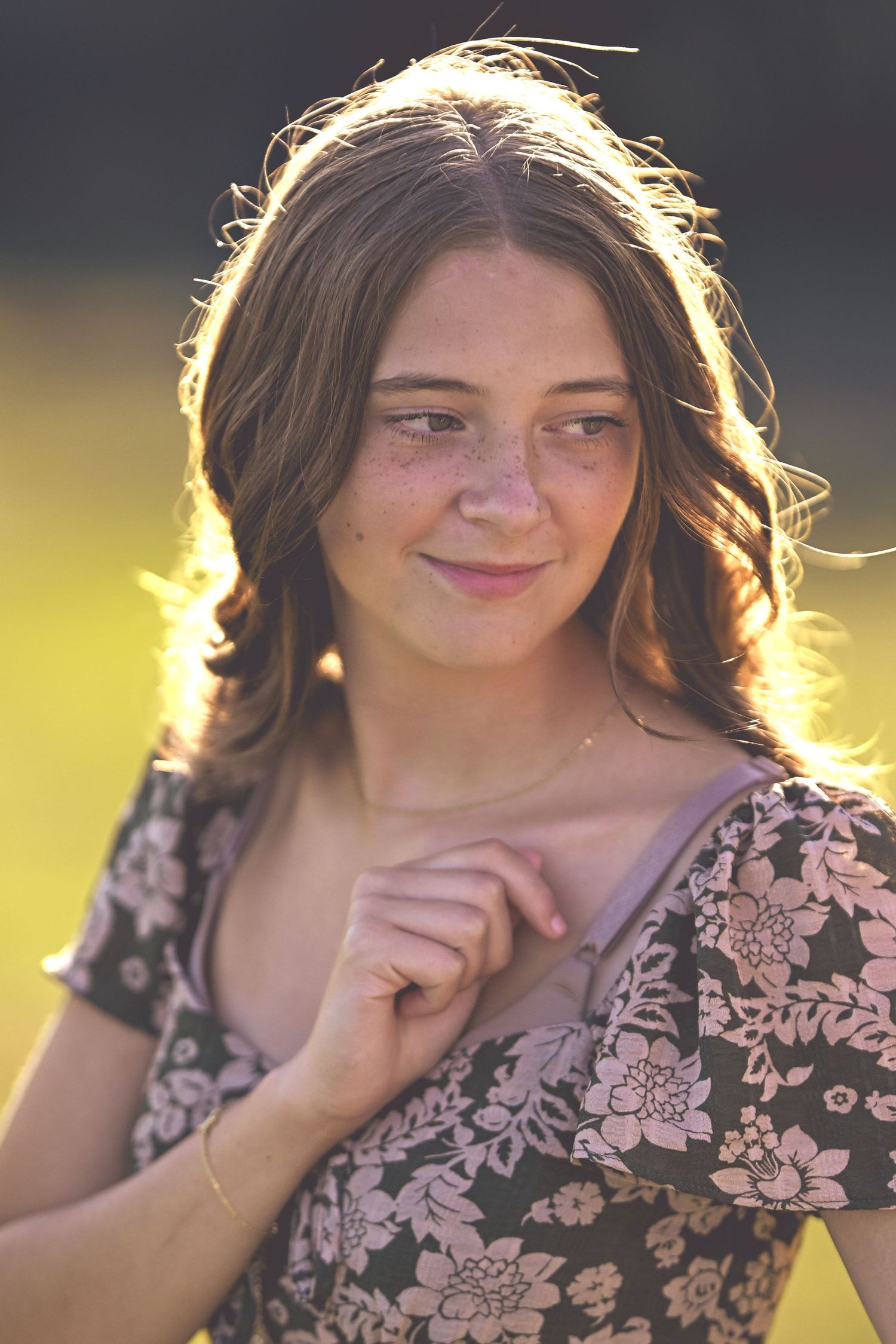 A person in a floral dress smiles warmly while backlit by golden sunlight in an outdoor portrait.