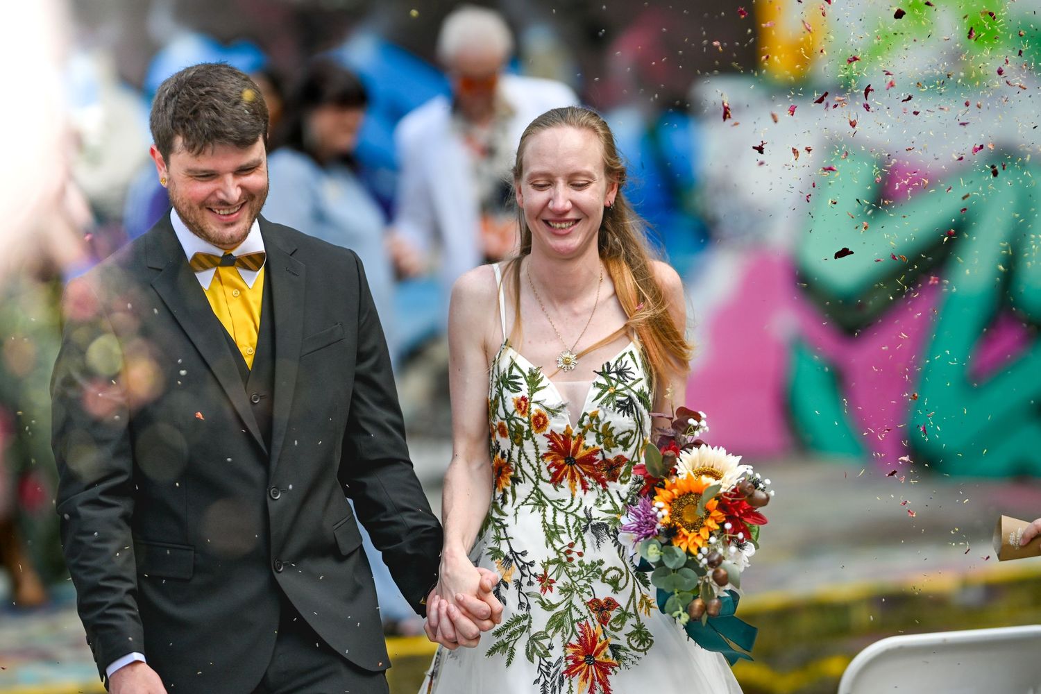 Newlywed couple walks outdoors surrounded by colorful confetti during joyful wedding celebration.